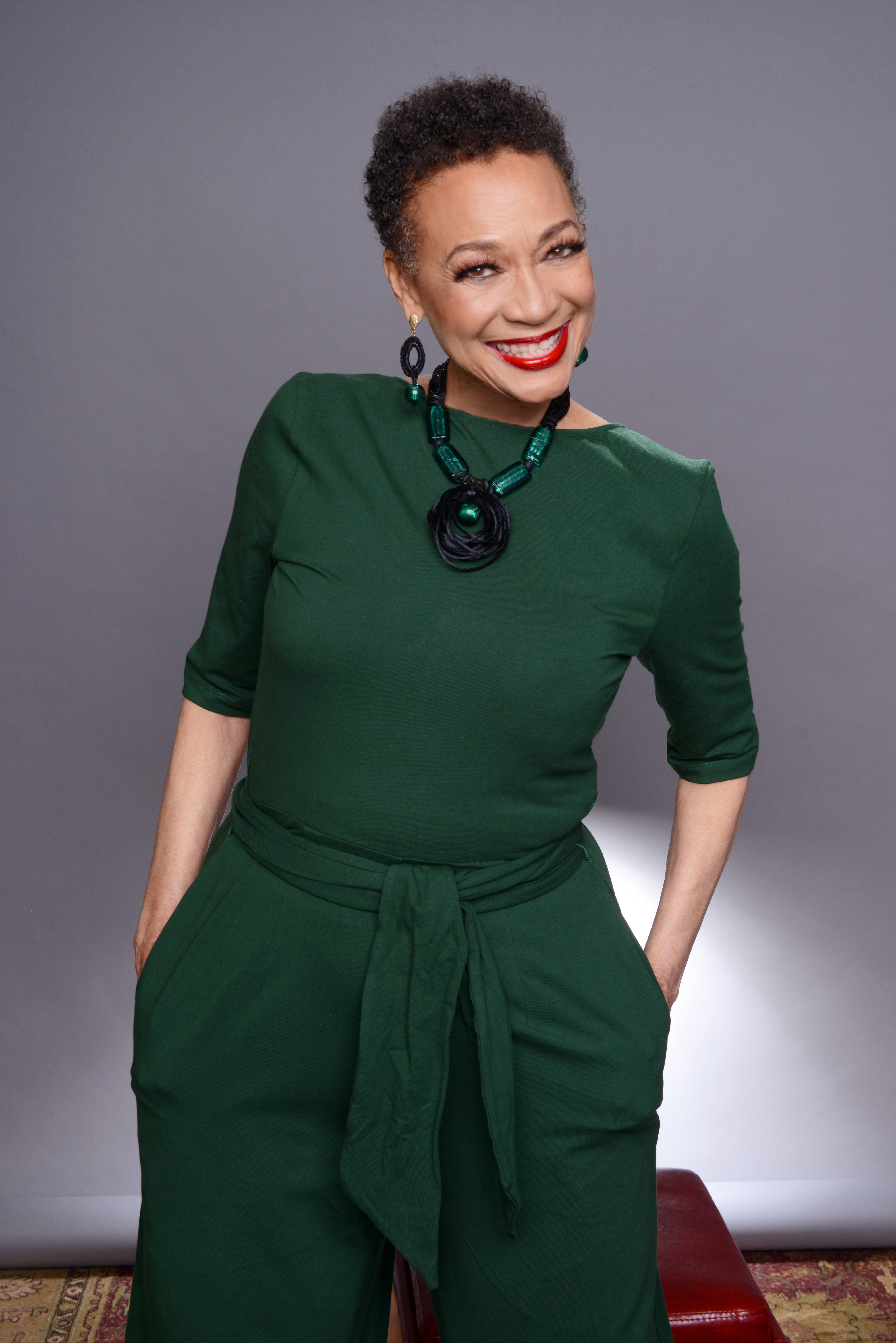Woman with short curly hair wearing a green dress and jewelry, smiling, standing against a gray background.