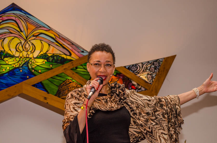 Person holding microphone, smiling, wearing animal print shawl, with colorful artwork in background.