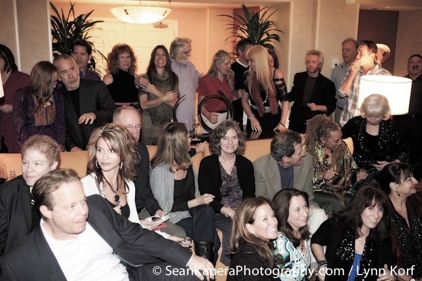 Group of people gathered indoors, some sitting and some standing, in a social setting with warm lighting and plants.