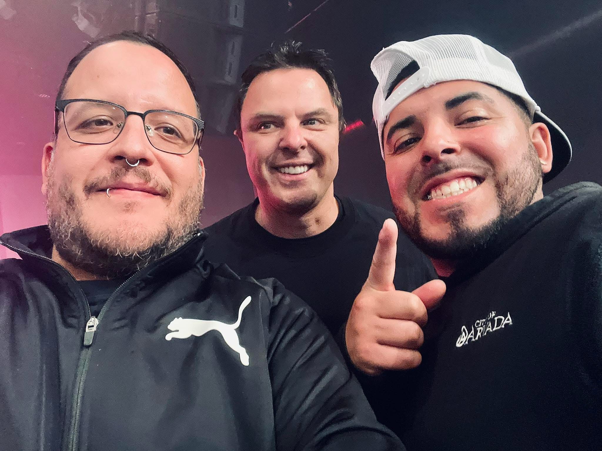 Three men smiling, one pointing, in a dark indoor setting, casual clothing, one with glasses and a cap.