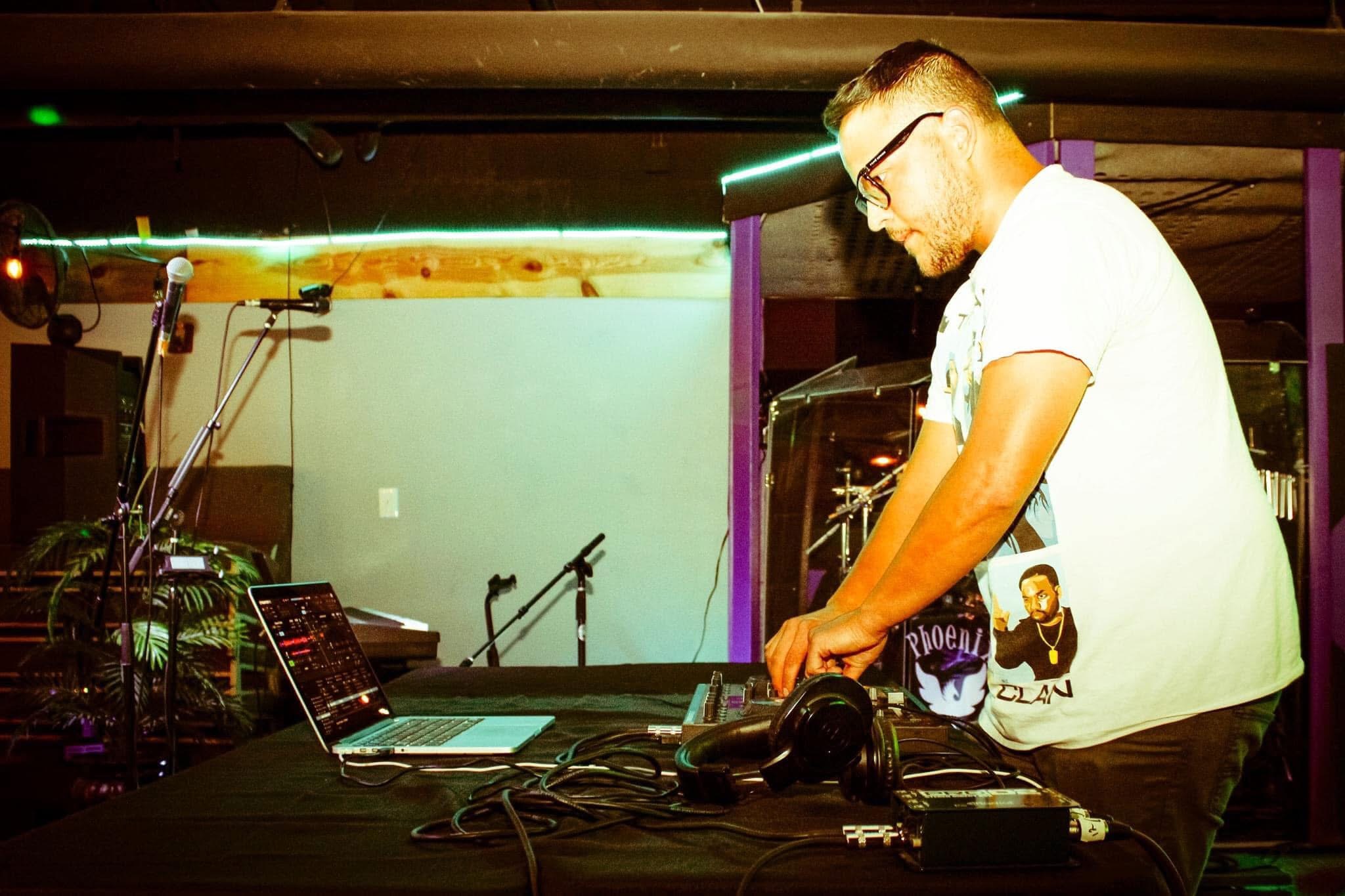 A man with glasses and a white T-shirt is DJing with equipment on a table, in a dimly lit venue.