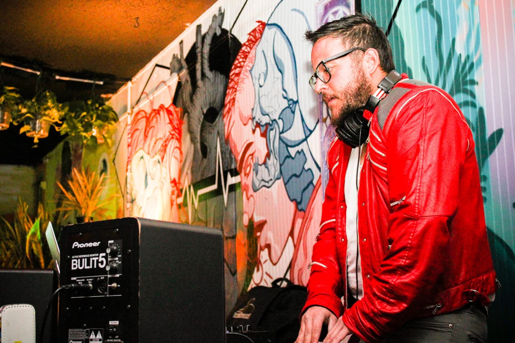 A man with glasses and a beard wearing a red jacket and headphones stands near DJ equipment in front of colorful graffiti art wall.