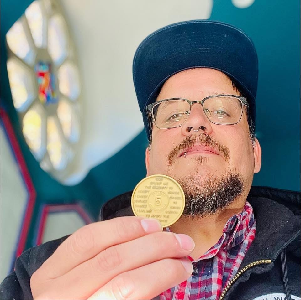 Person wearing glasses and a cap holding a gold-colored coin, smiling slightly, with a colorful background.