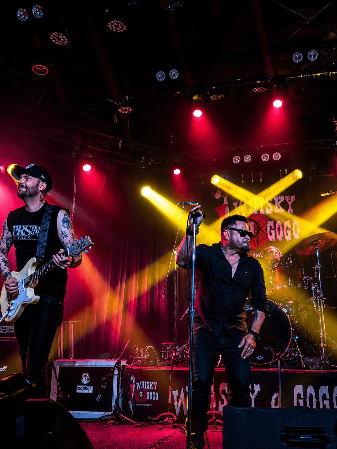Two musicians perform on stage with colorful lights and a banner in the background, one singing into a microphone and the other playing guitar.