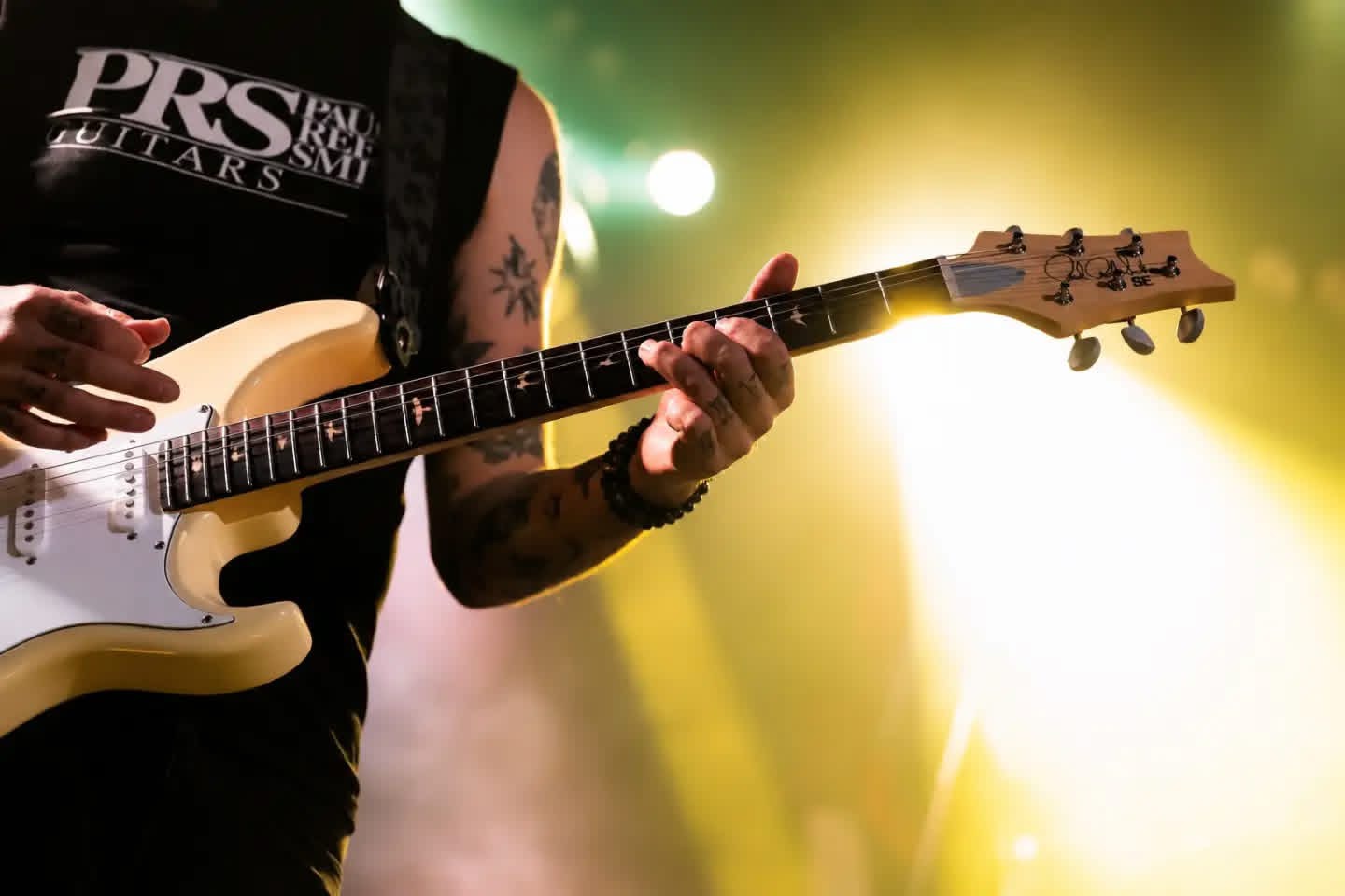 Person playing electric guitar on stage with bright yellow and green lighting, tattoos visible on arm, wearing a sleeveless shirt.