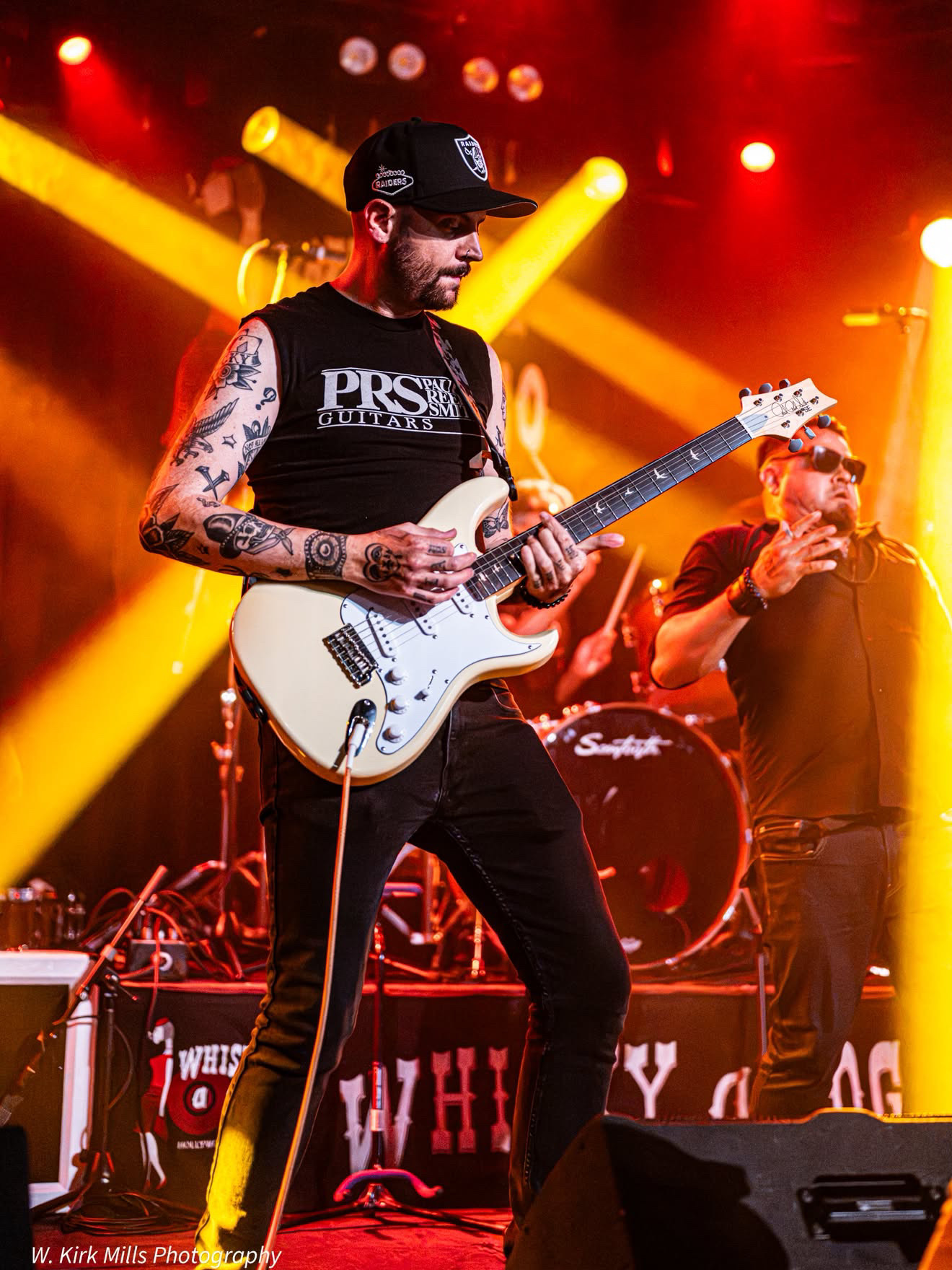Musician playing white electric guitar on stage with yellow and red lights, wearing a black cap and sleeveless shirt, tattoos visible