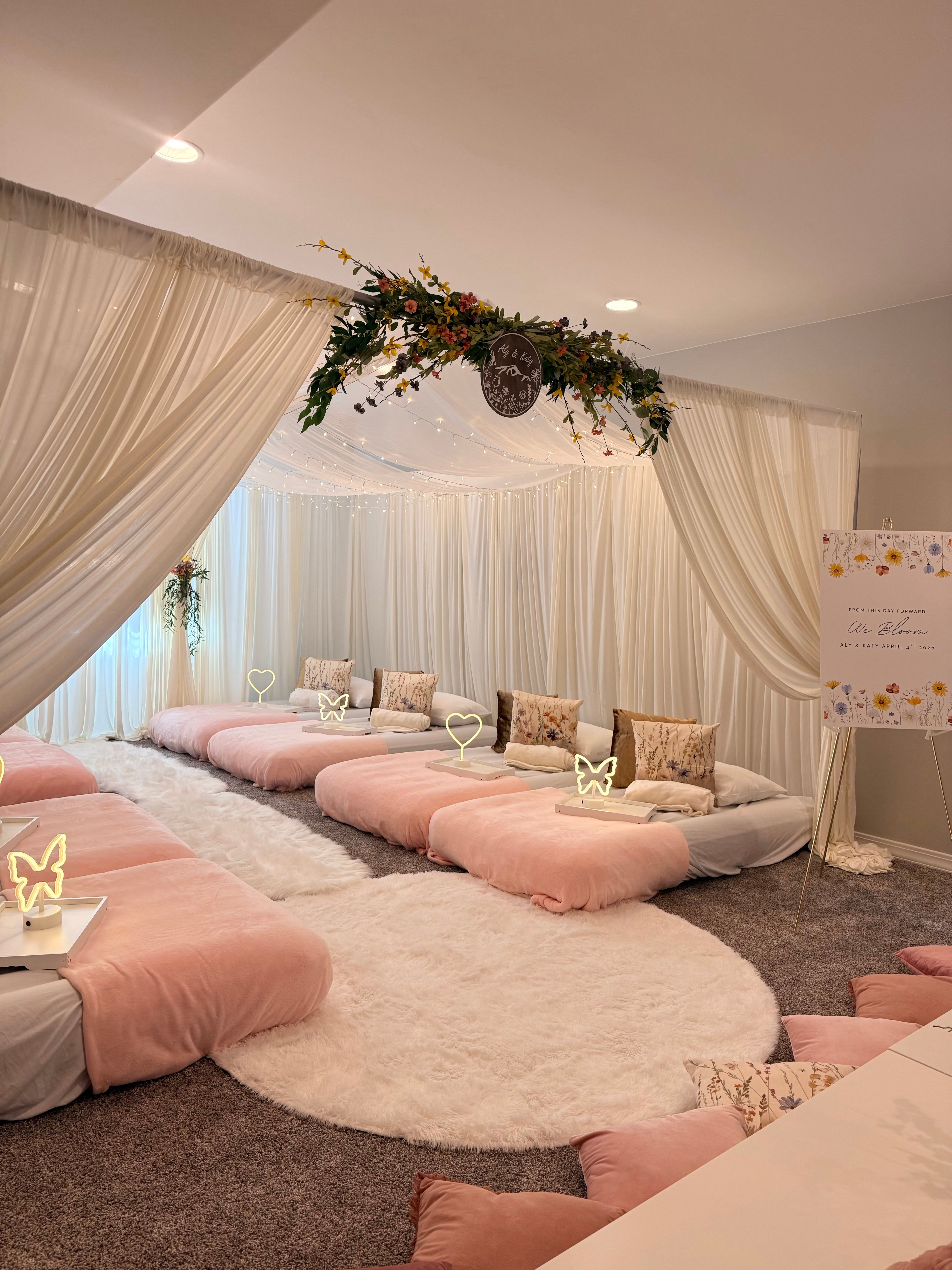 Indoor event space with pink cushions, decorative pillows, a white circular rug, and floral hanging decoration, curtains, and a whiteboard.