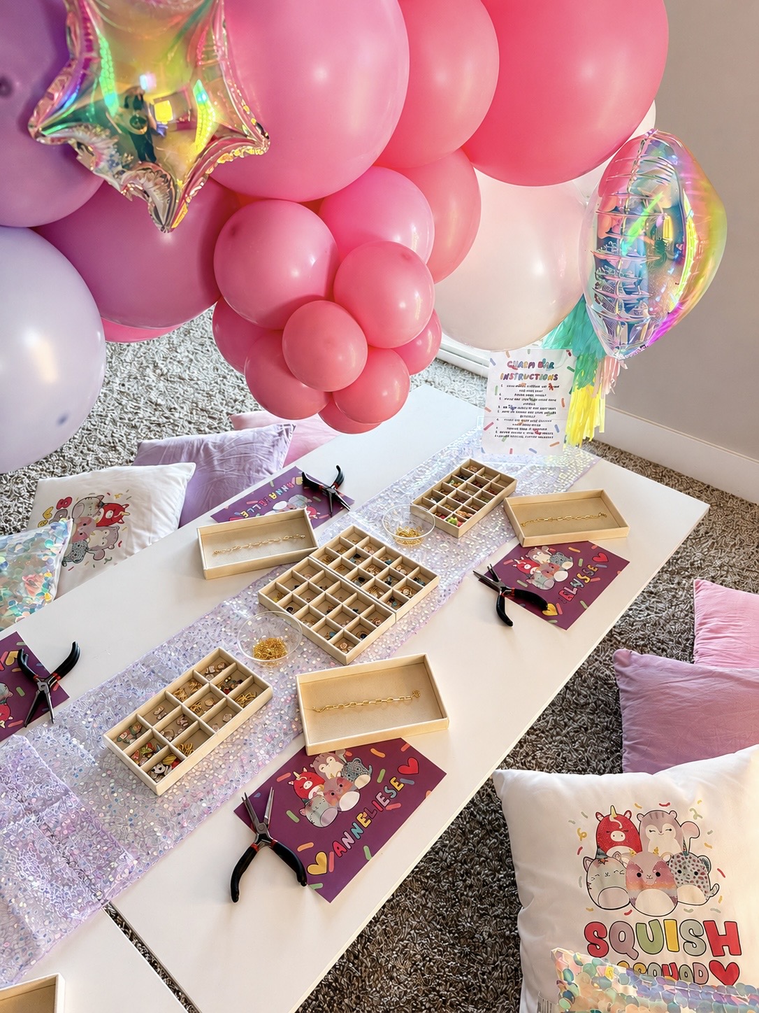 Party table with balloons, chocolates, and gift boxes, decorated with pink, purple, and holographic balloons, and pillows nearby.