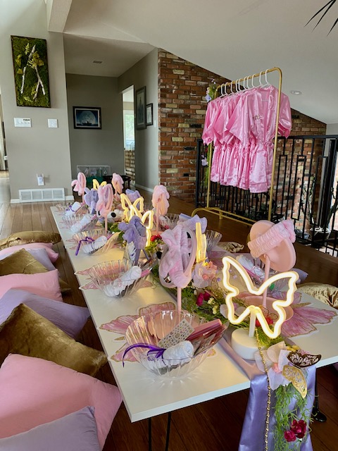 Decorated table with pink and purple items, butterfly lights, and pink dresses hanging nearby in a room with brick wall.