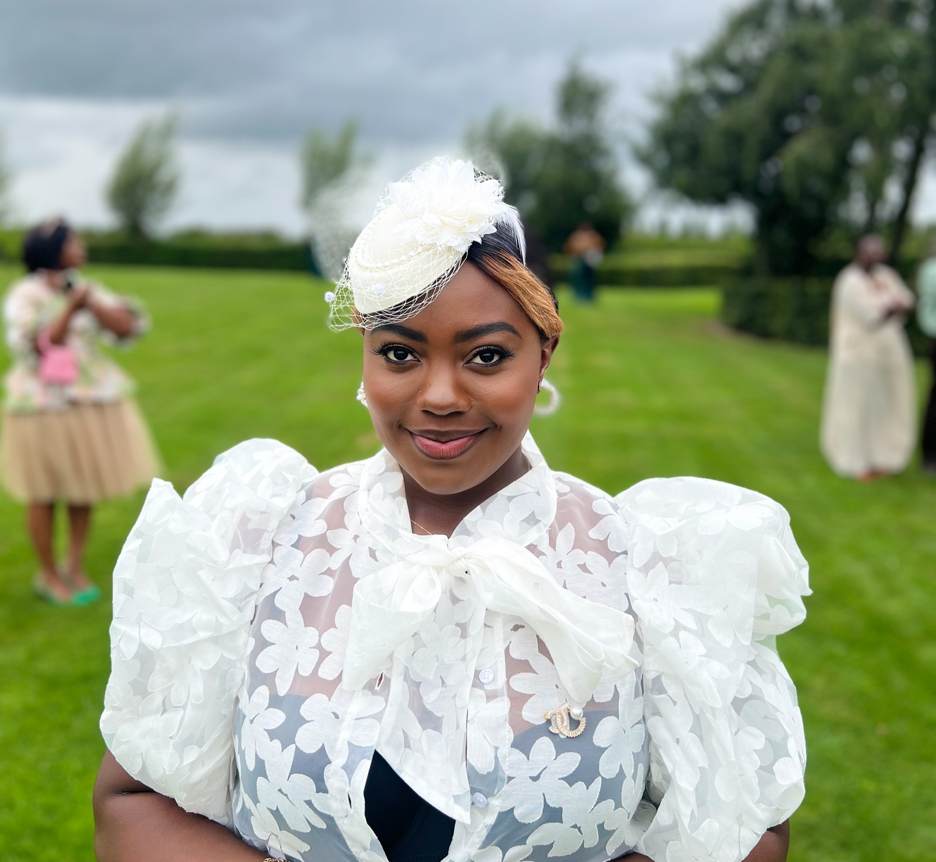 Young woman outdoors wearing a white lace dress with puffed sleeves and a headpiece, smiling at the camera.