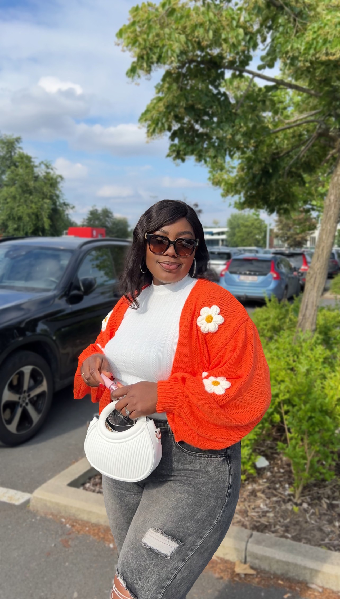 Woman wearing sunglasses and a bright orange jacket with white flowers, holding a white bag, standing outdoors near parked cars and trees.