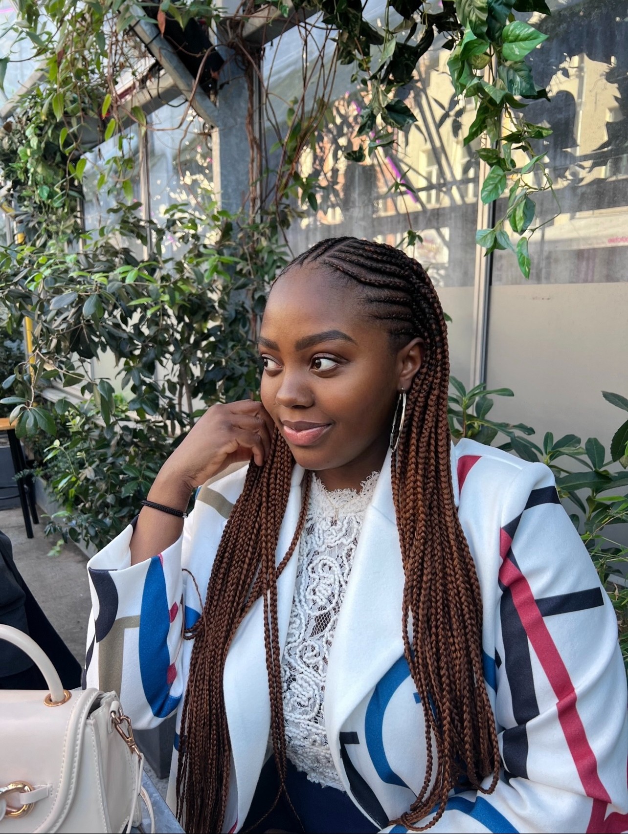 Young woman with braided hair sitting outdoors among greenery, wearing a white jacket with colorful stripes and lace top.