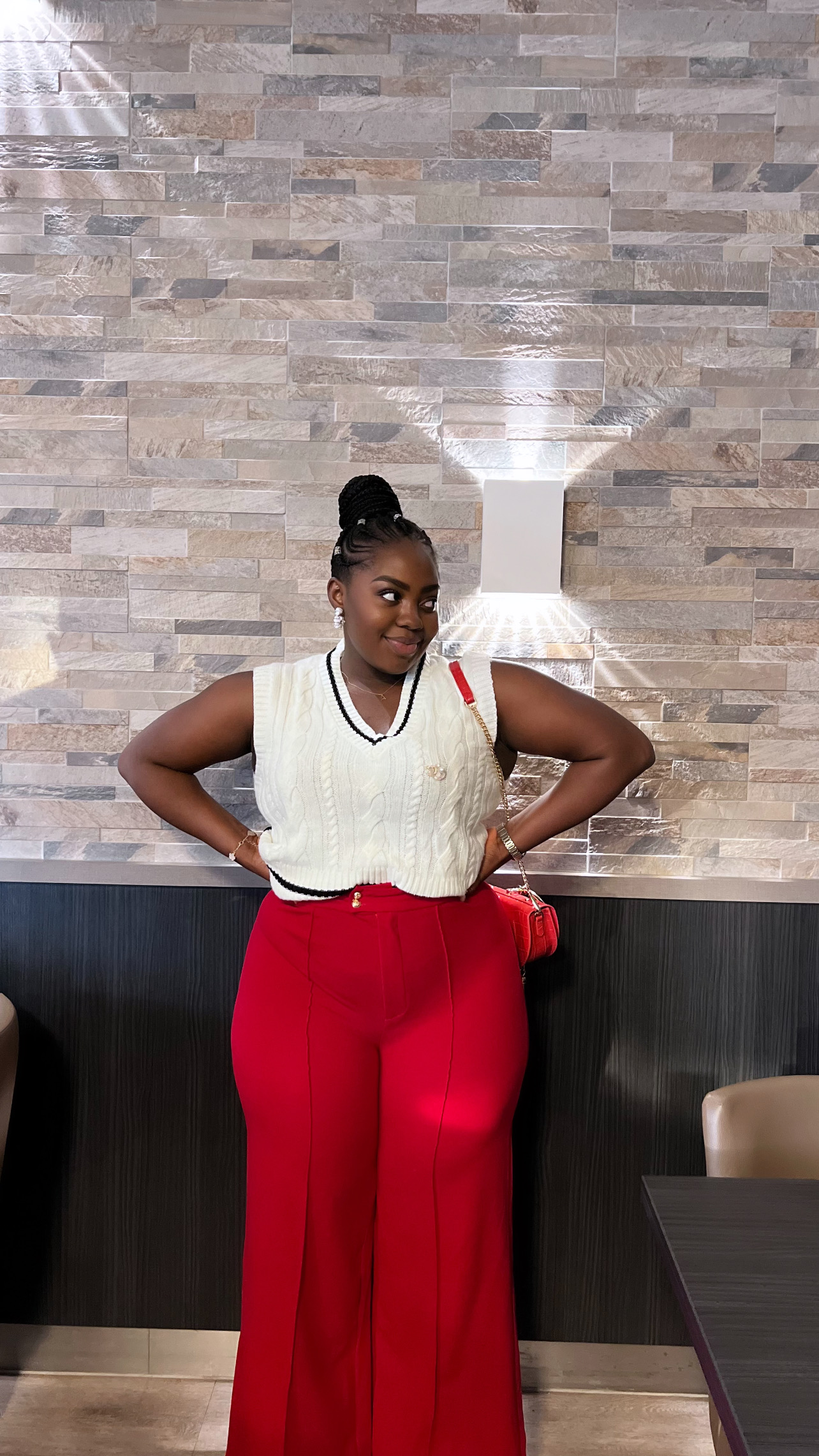 Woman standing with hands on hips in front of a stone wall and white vase, wearing a white top and red pants.