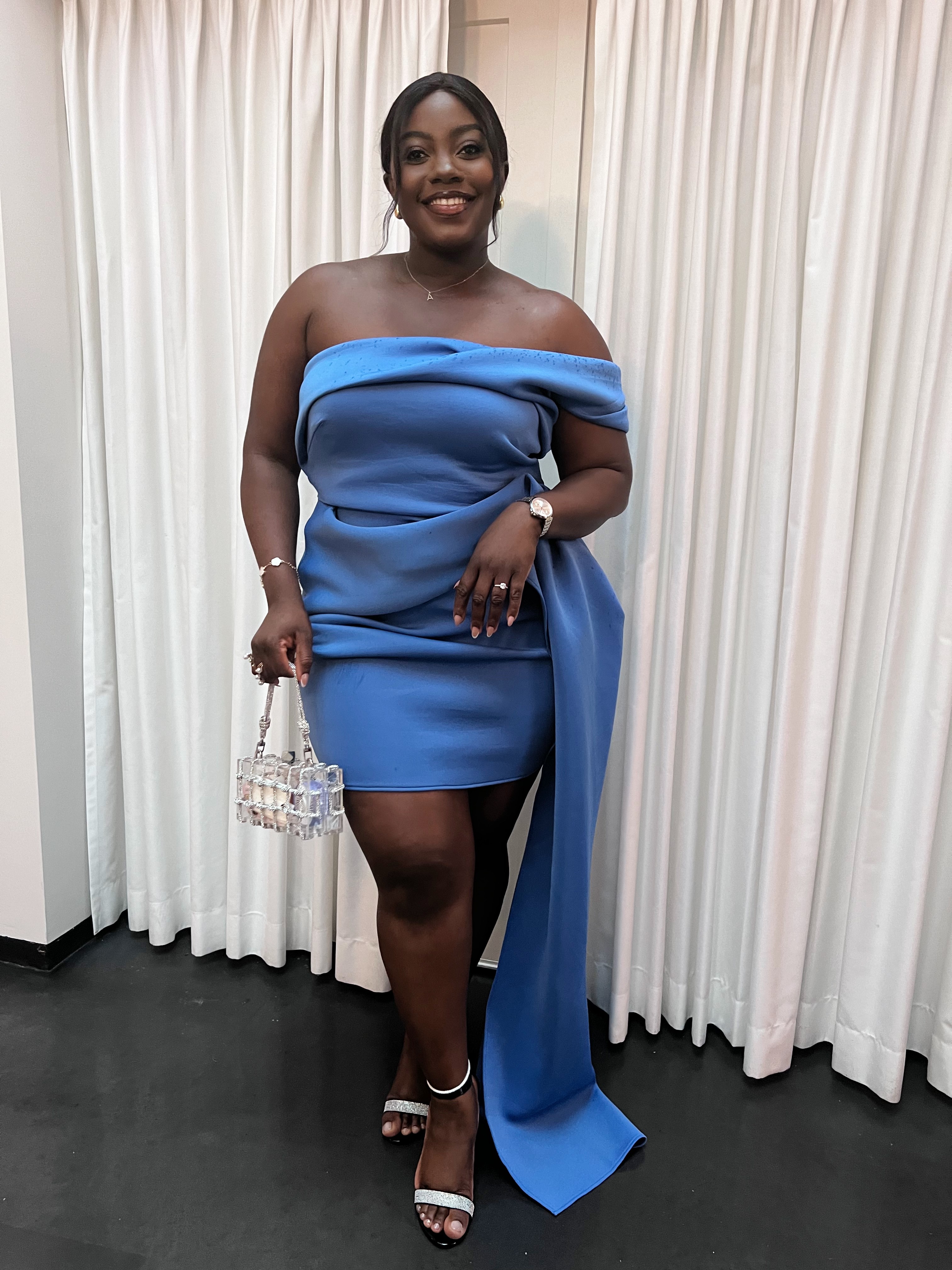 Woman in blue off-shoulder dress holding a small handbag, standing in front of white curtains, smiling.