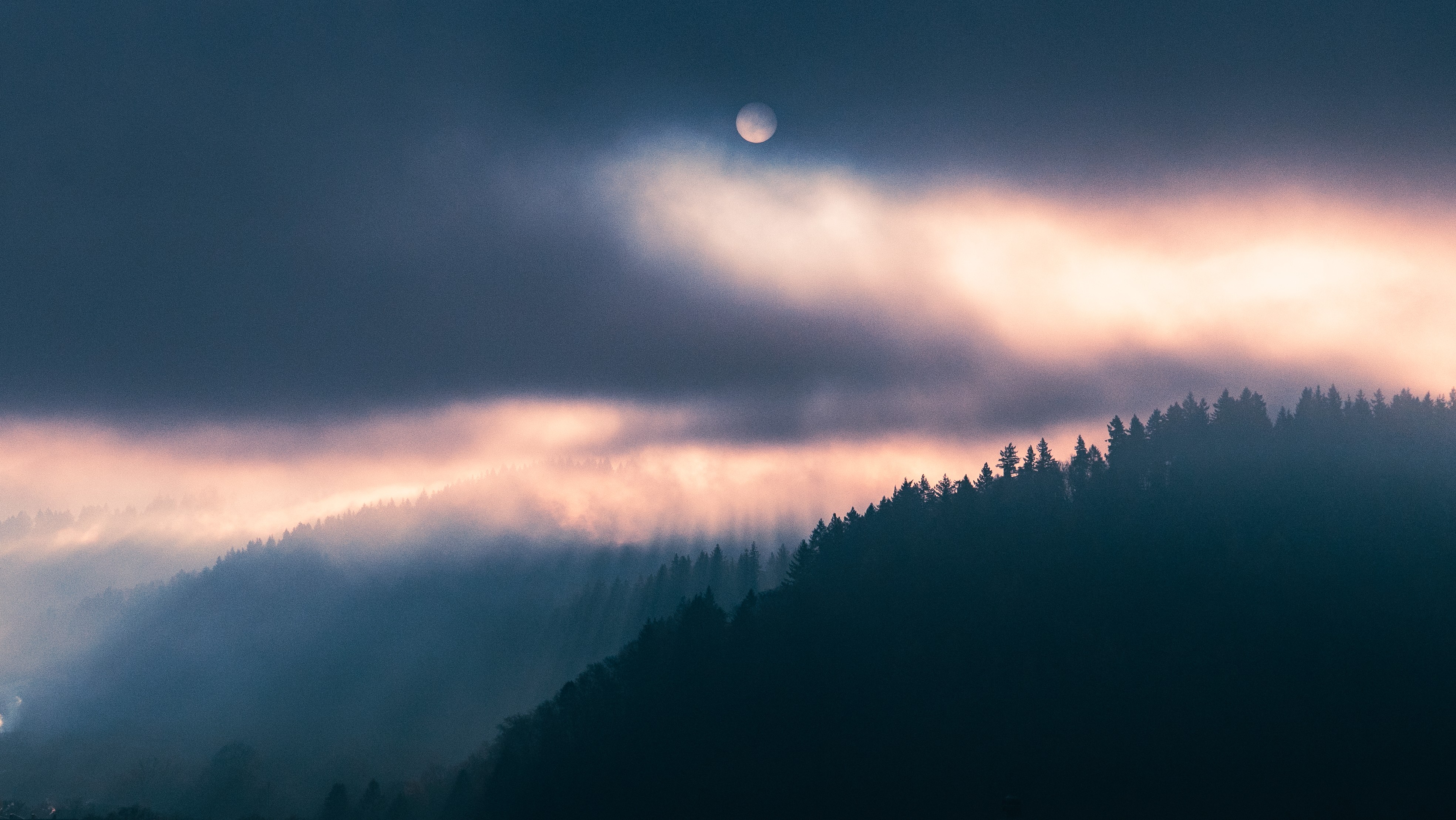 Nighttime sky with moon partially obscured by dark clouds over a forested mountain silhouette.