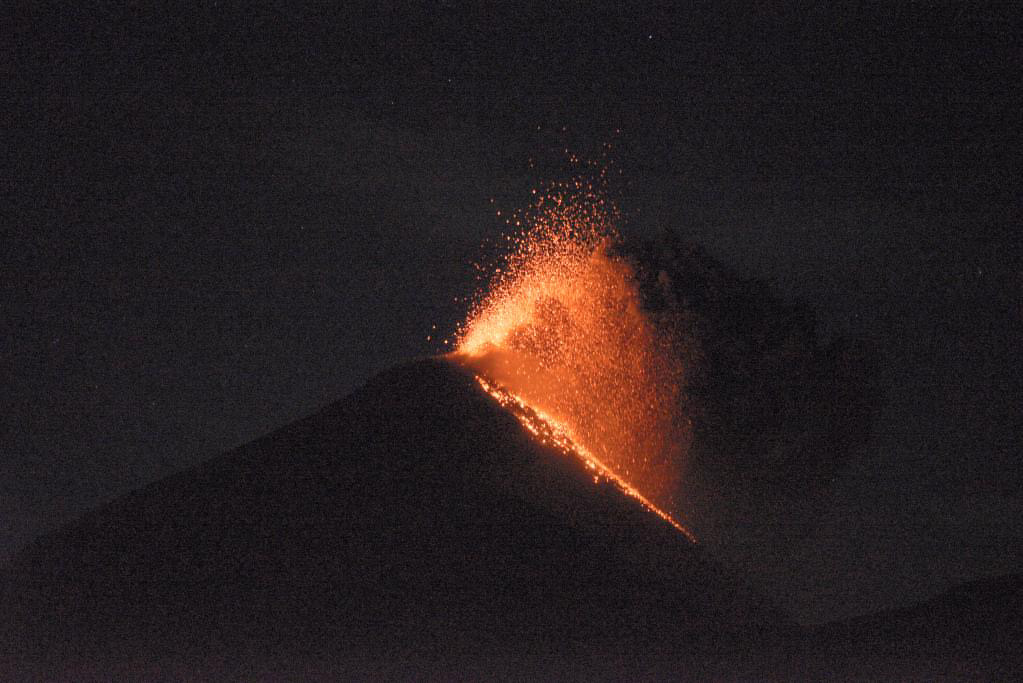 Eruption of lava and ash from a volcano at night, with glowing lava flowing down the side.