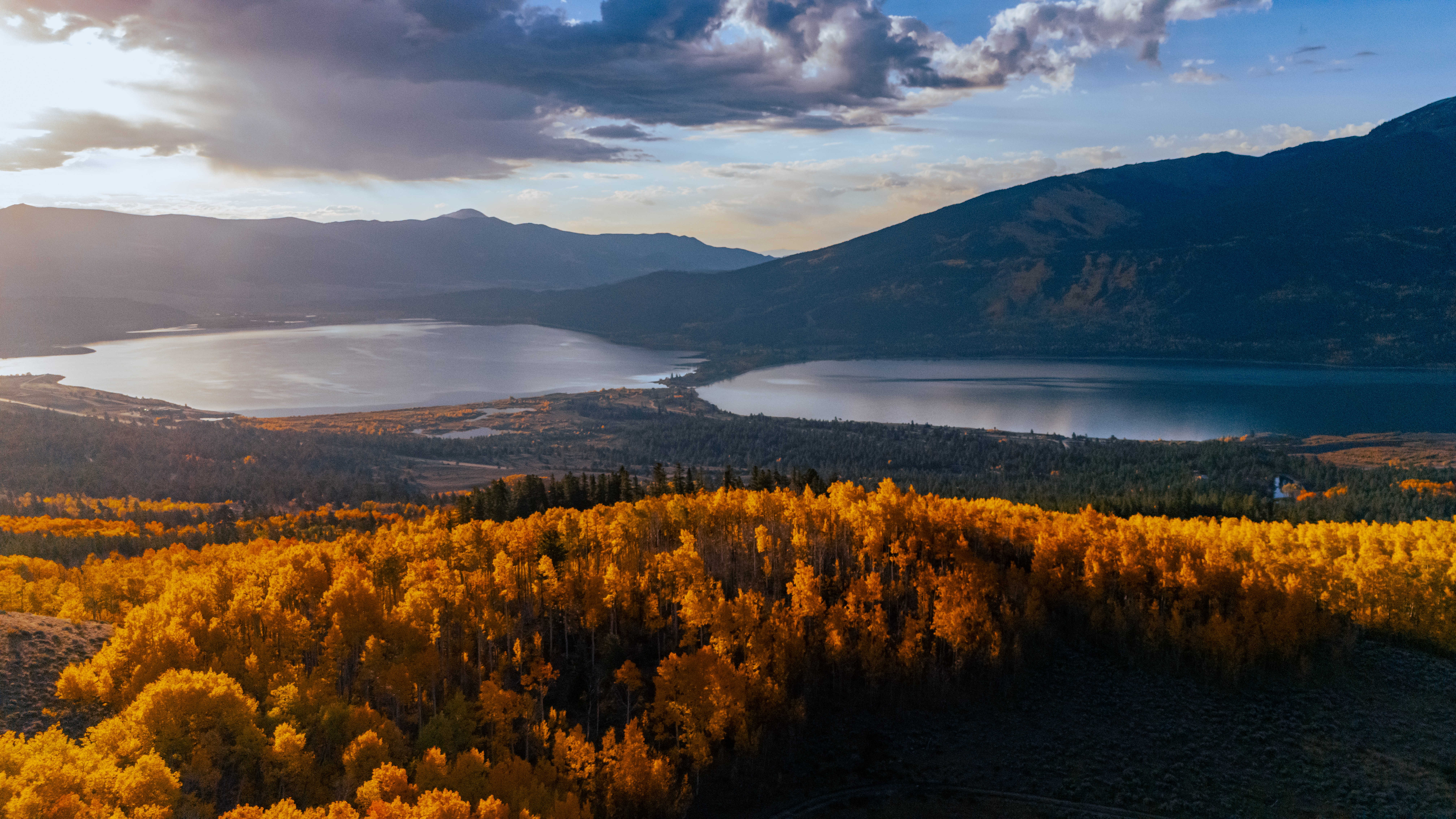 Landscape with a lake, mountains, and a forest with autumn-colored trees under a partly cloudy sky.