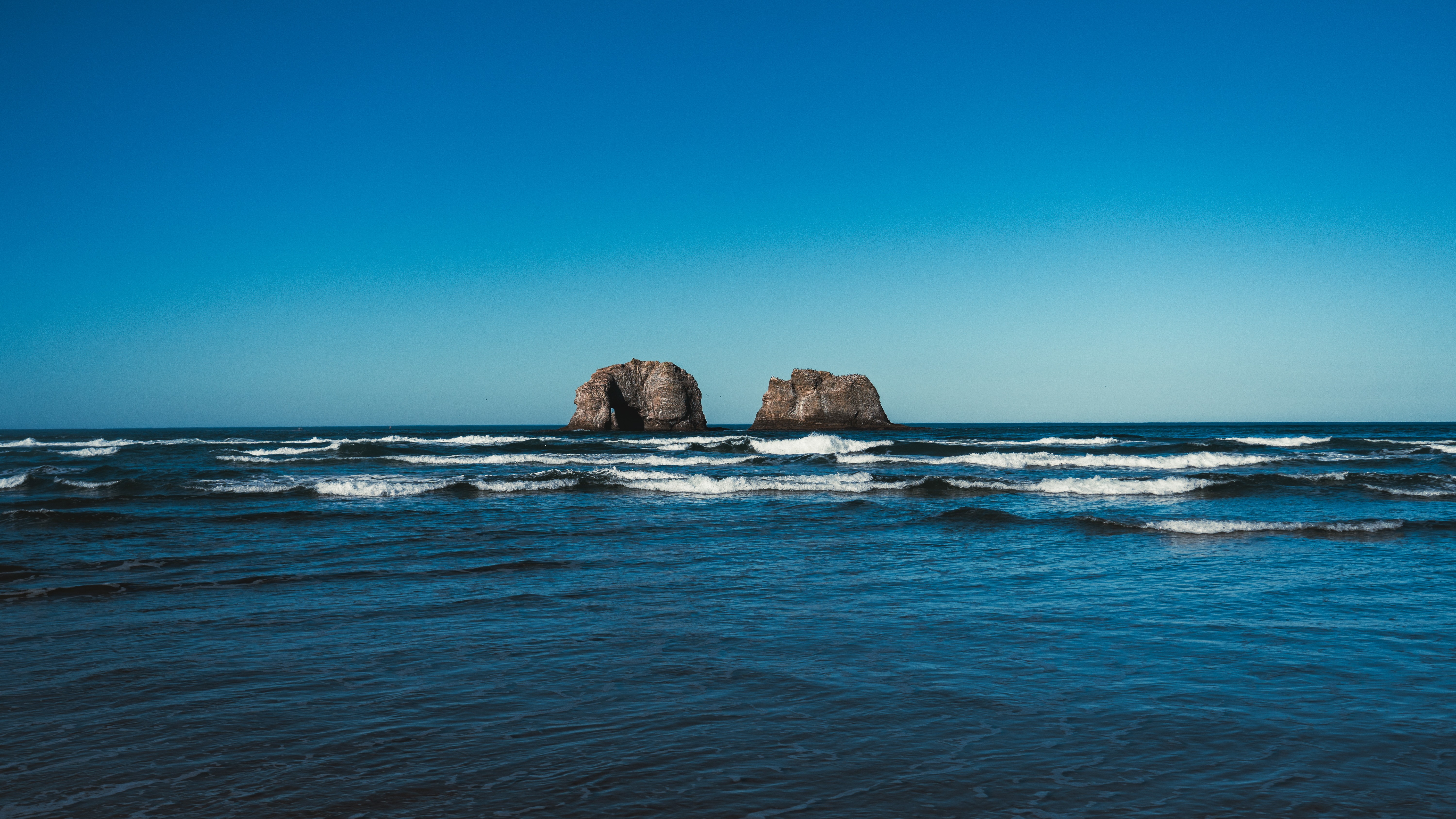 Ocean with waves and two large rocks in the distance under a clear blue sky.