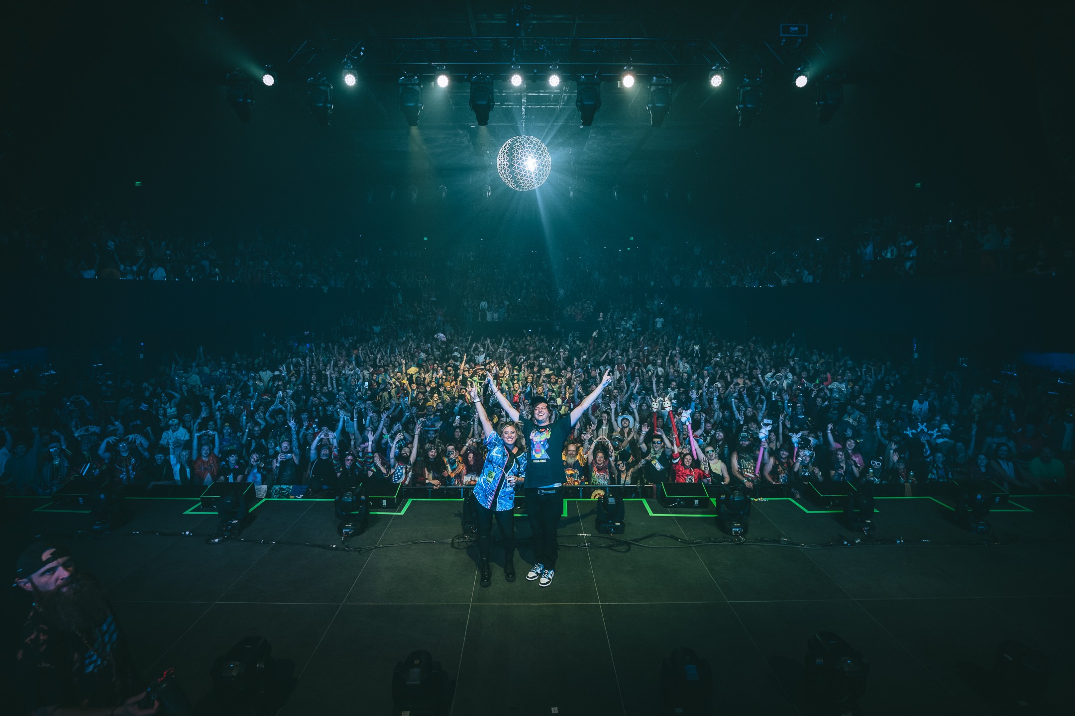 Person on stage with arms raised in front of a large audience under stage lights, with a disco ball overhead.