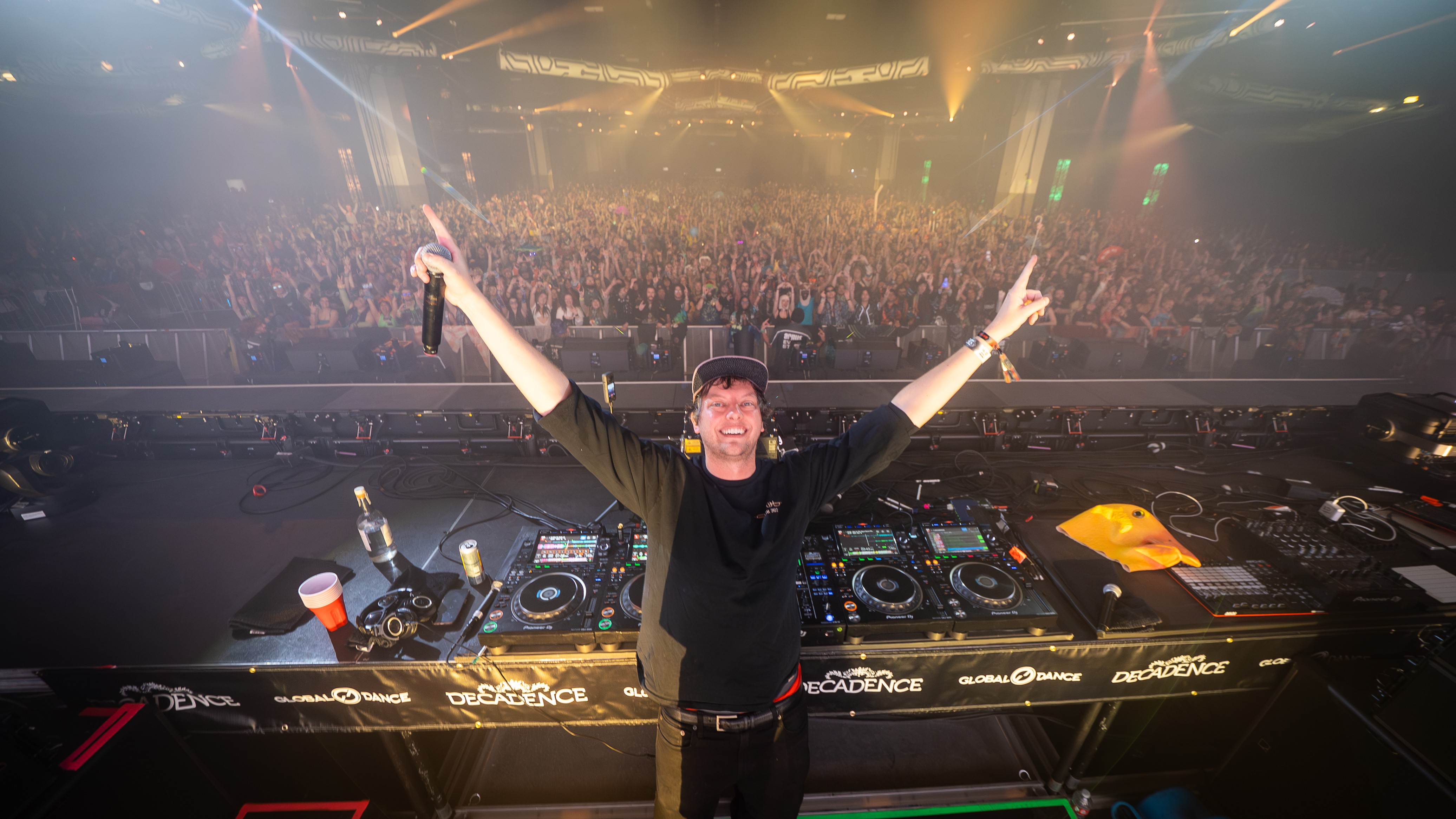 DJ standing with arms raised in front of a large crowd at a concert or festival, with DJ equipment and colorful lighting.