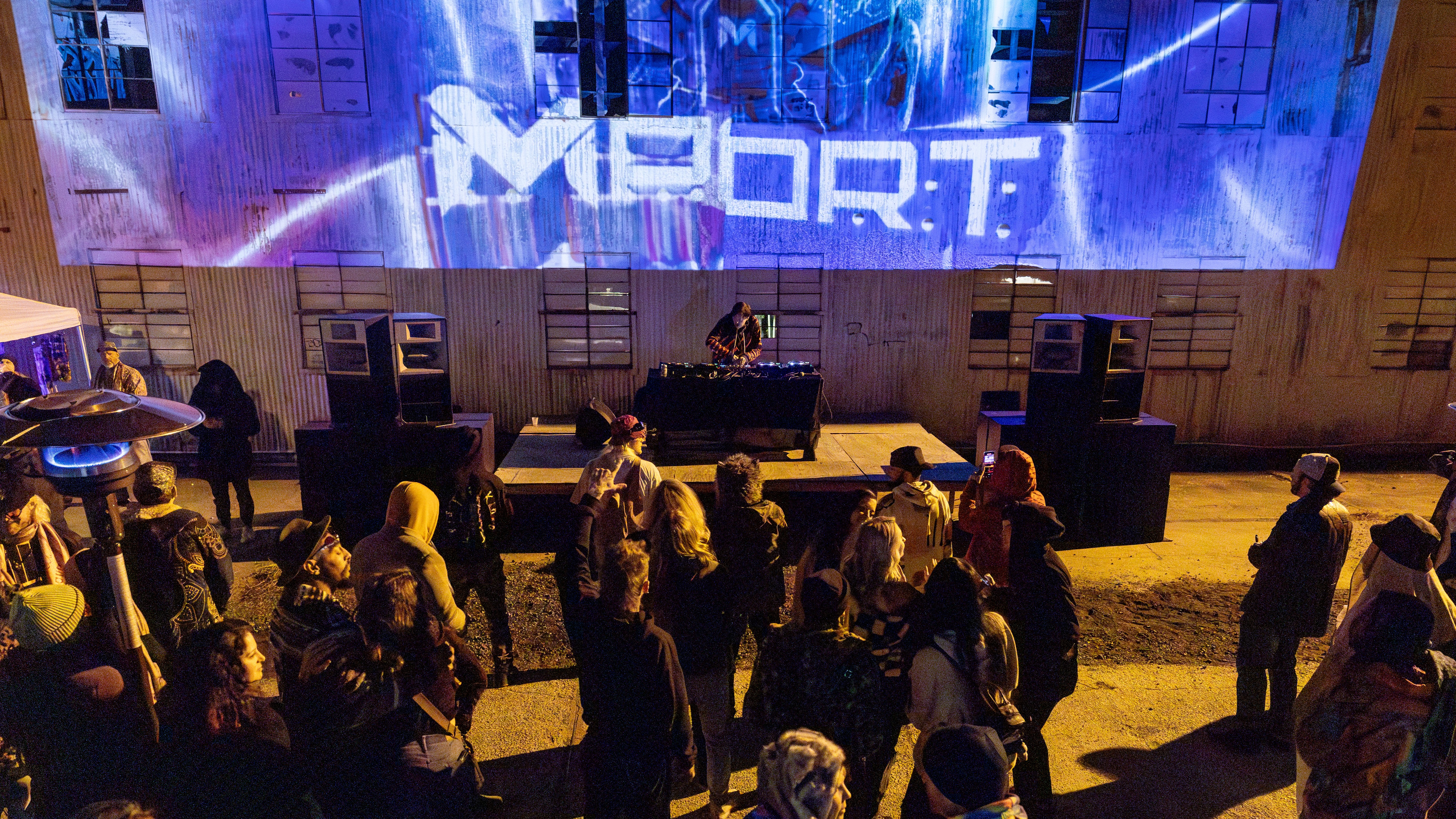 People gather outdoors at night watching a DJ perform on stage with a large screen displaying 'MYORT' behind them.