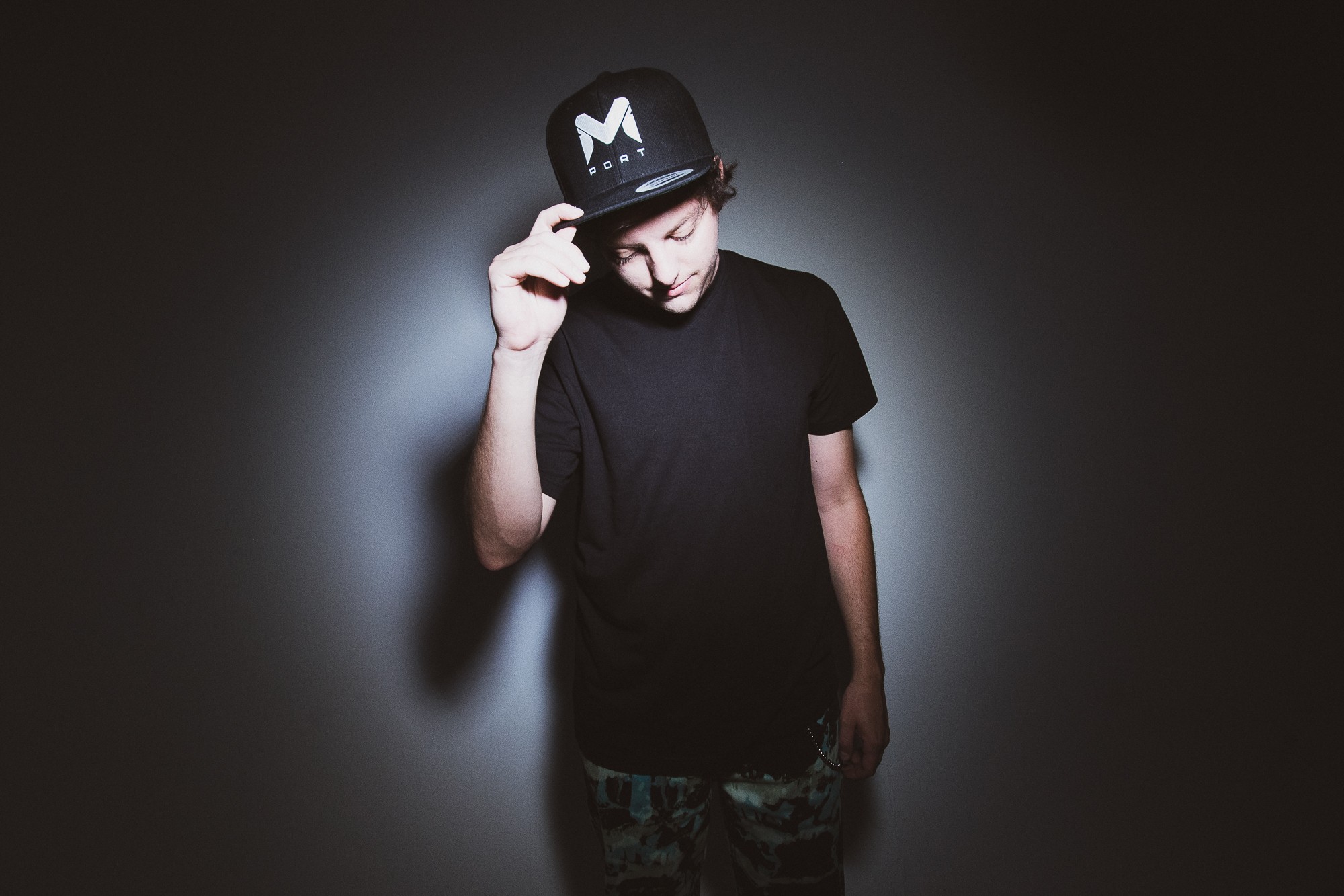 Young person wearing a black cap and t-shirt, standing against a dark background with a light halo, looking down.