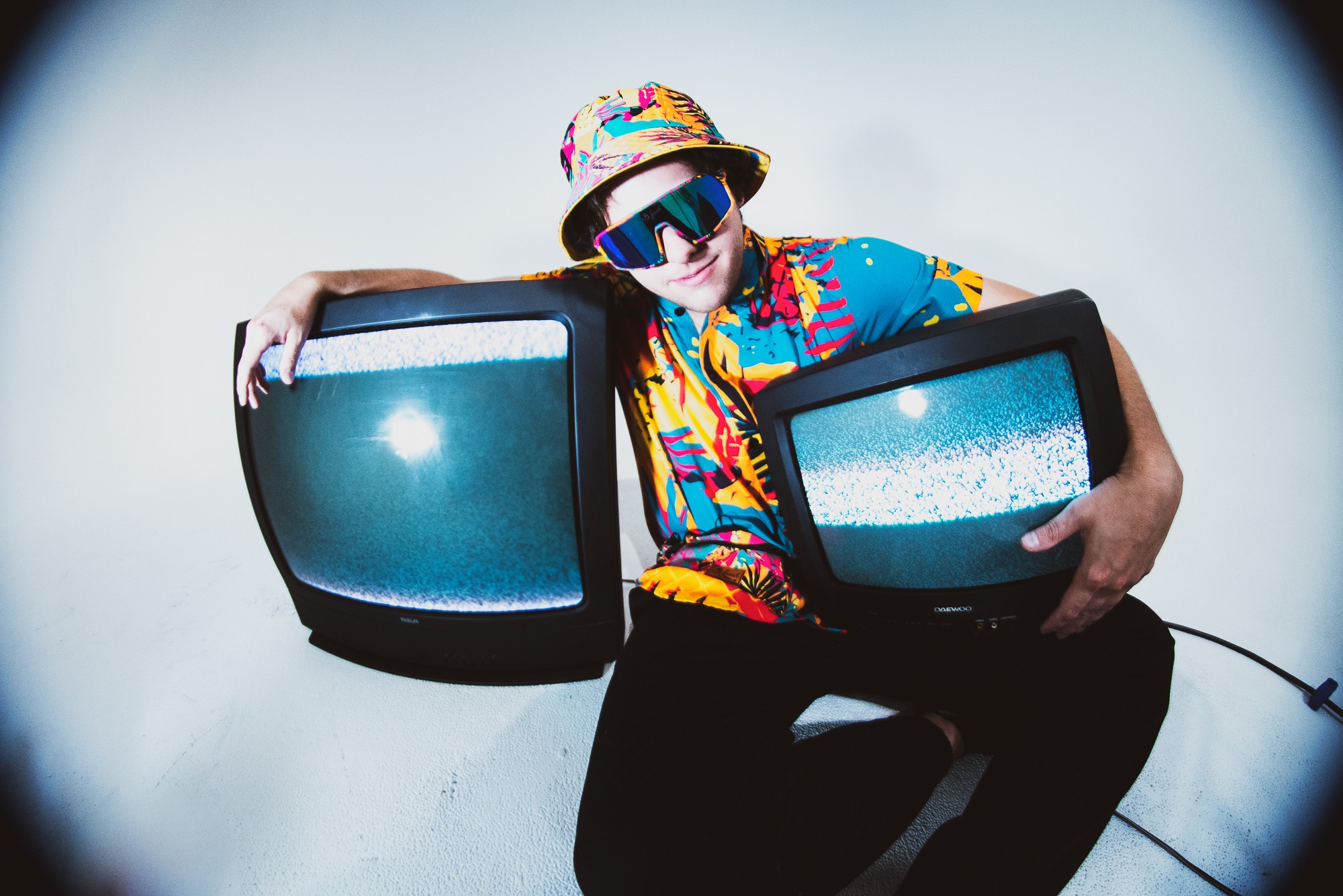 Person wearing colorful clothing, sunglasses, and a hat, holding two vintage televisions, against a plain background.