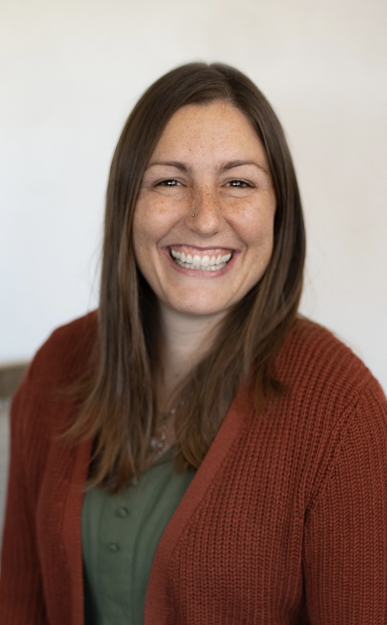 Smiling woman with long brown hair wearing a green top and rust-colored cardigan, against a plain background.