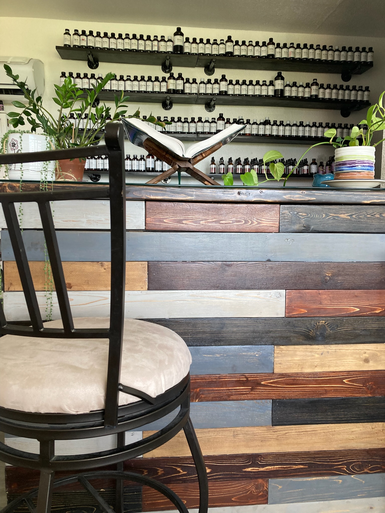 Indoor space with a wooden counter, potted plants, and shelves with small bottles or containers, and a black chair with a cushioned seat.