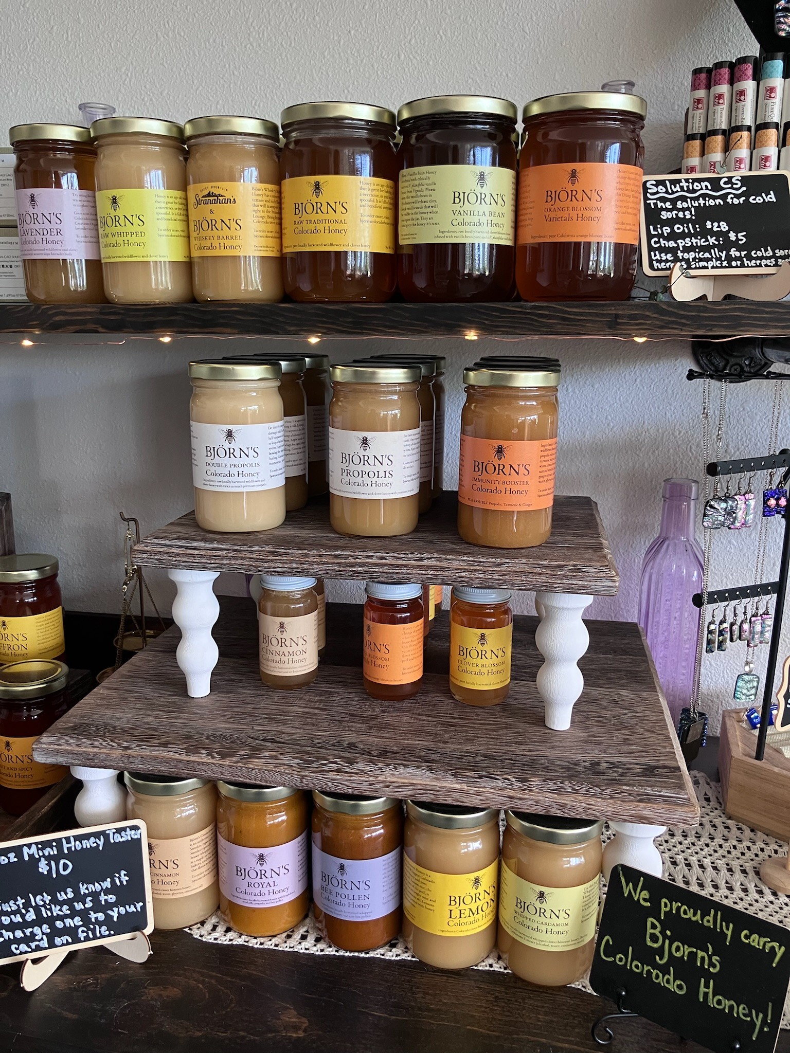Jars of honey and honey products displayed on shelves and a table, with a chalkboard sign and a purple container in the background.
