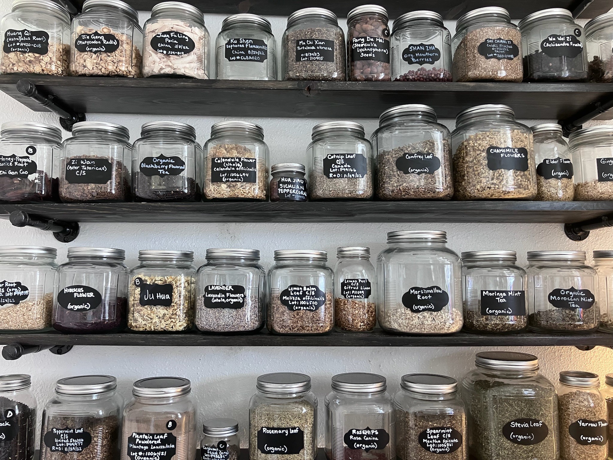 Multiple glass jars with black labels on shelves, containing various grains and spices, arranged in rows.