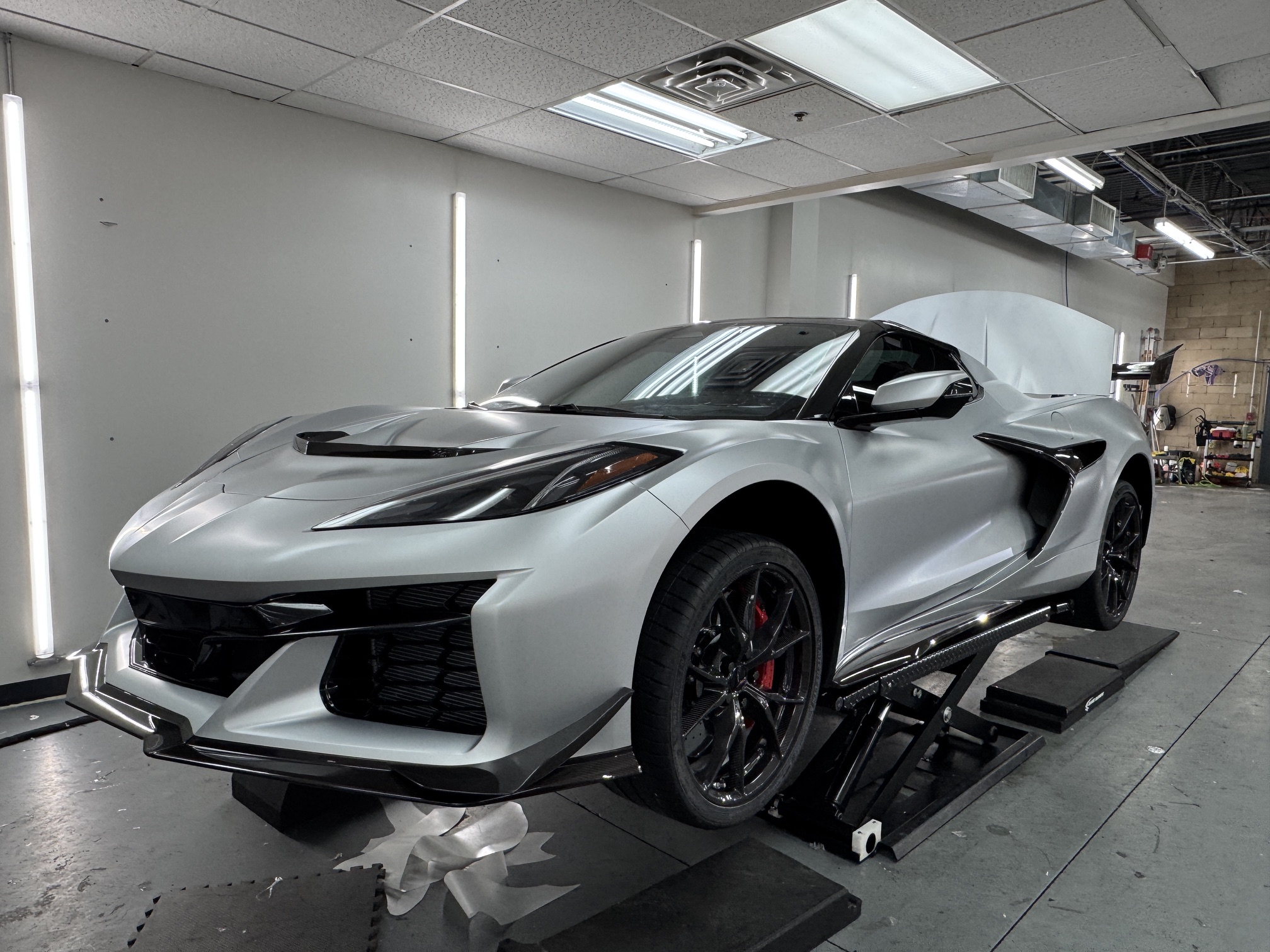 White sports car with black wheels and red brake calipers inside a garage, on a platform, with ceiling lights overhead.