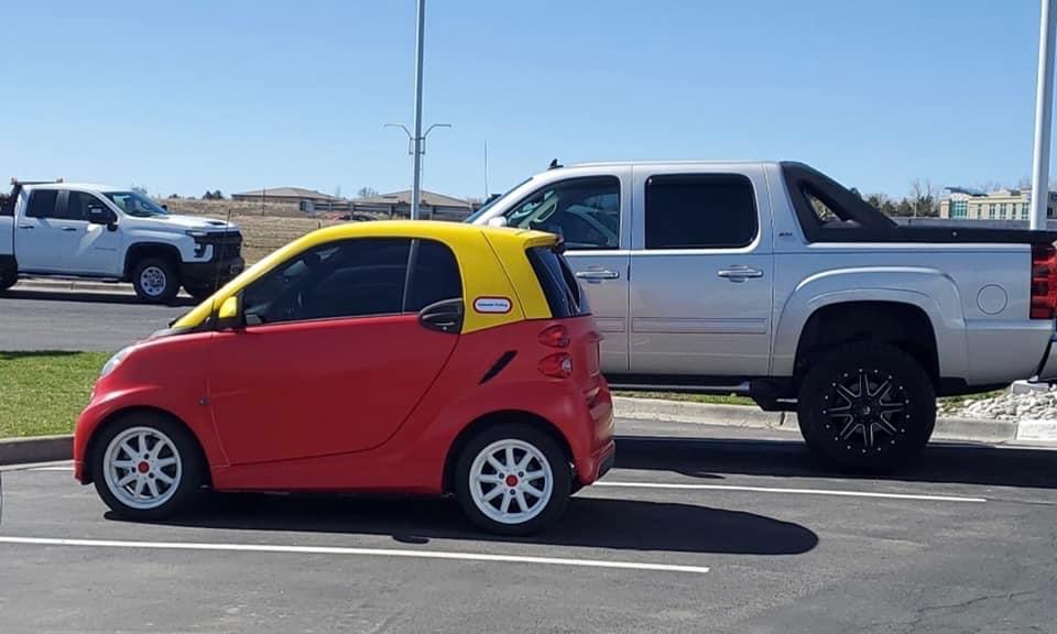 Small red and yellow compact car parked next to a large silver pickup truck in a parking lot.