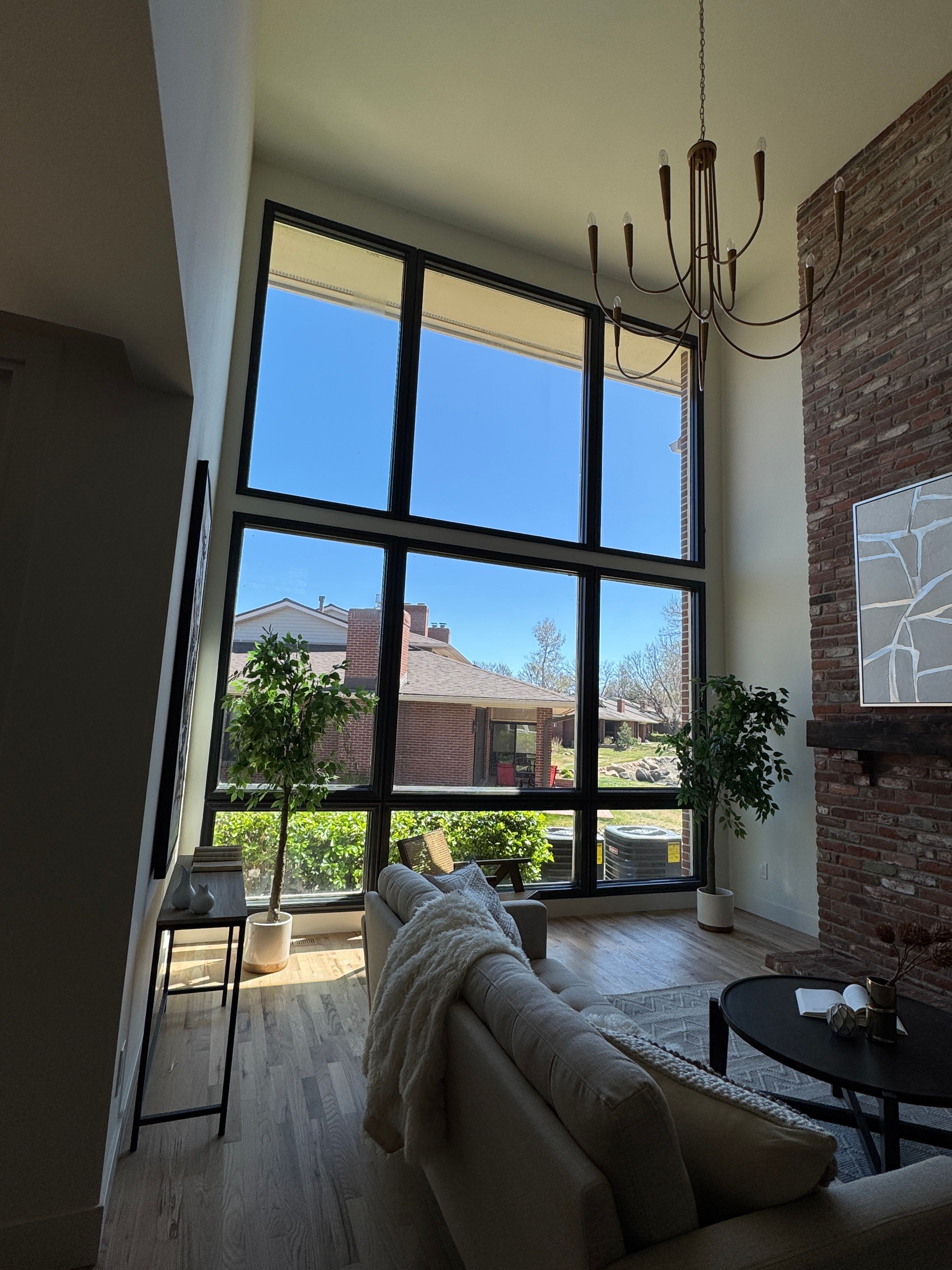 Living room with large window, plants, and furniture, viewed from an angle, with sunlight and outdoor scene visible.
