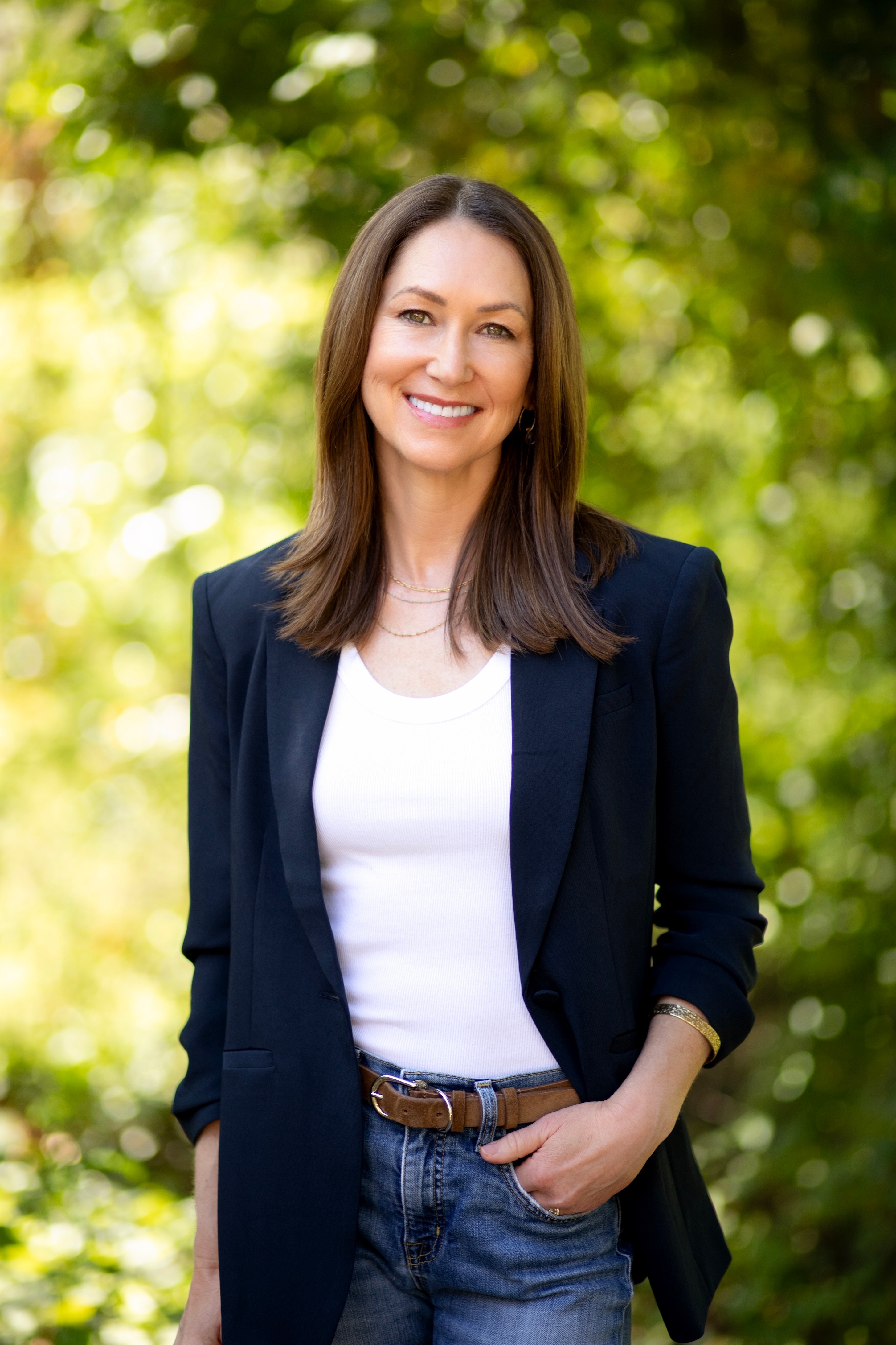 Woman with long brown hair smiling outdoors, wearing a dark blazer over a white shirt, with trees in the background.