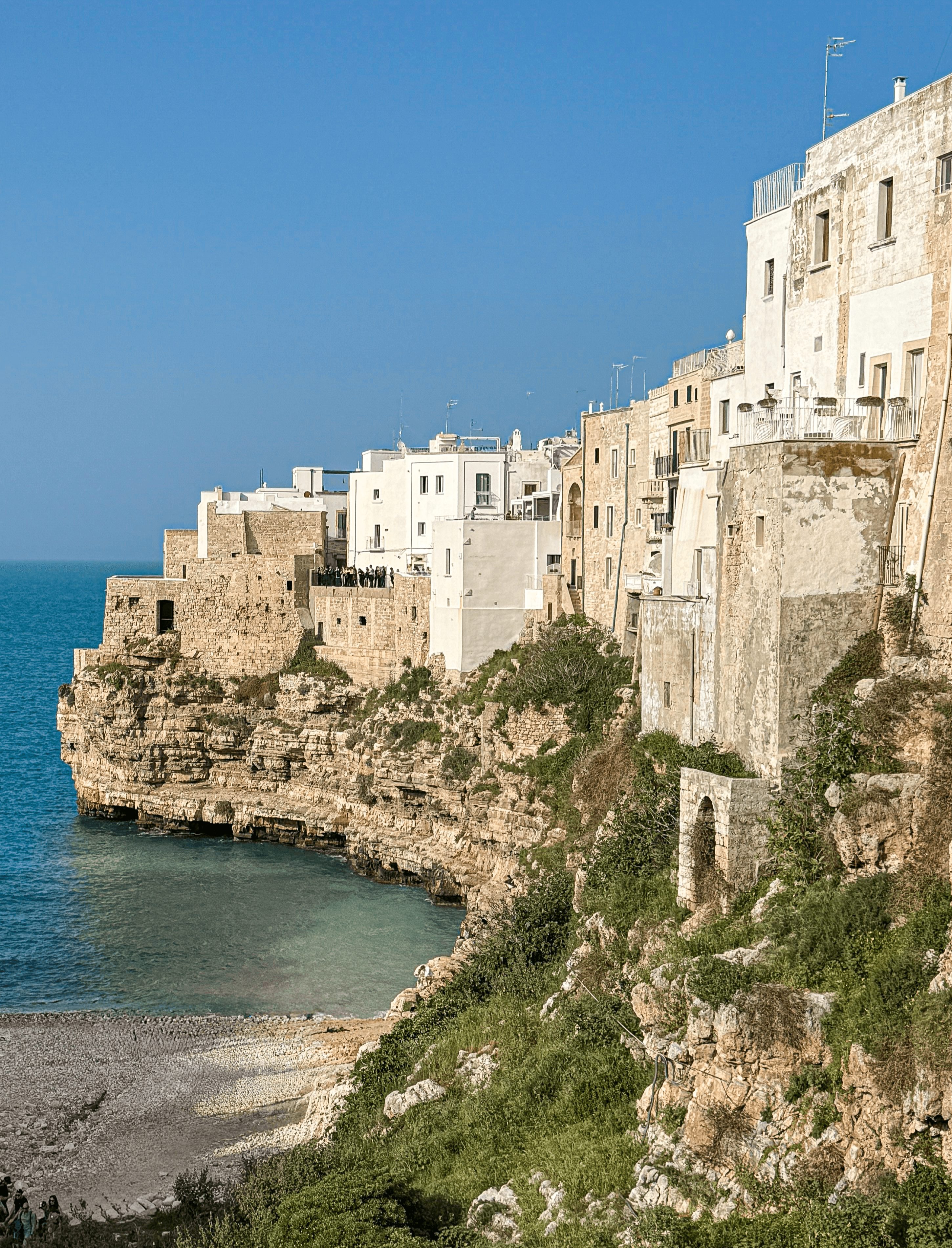 Cliffside buildings with white and beige facades overlooking a small beach and the sea under a clear blue sky.