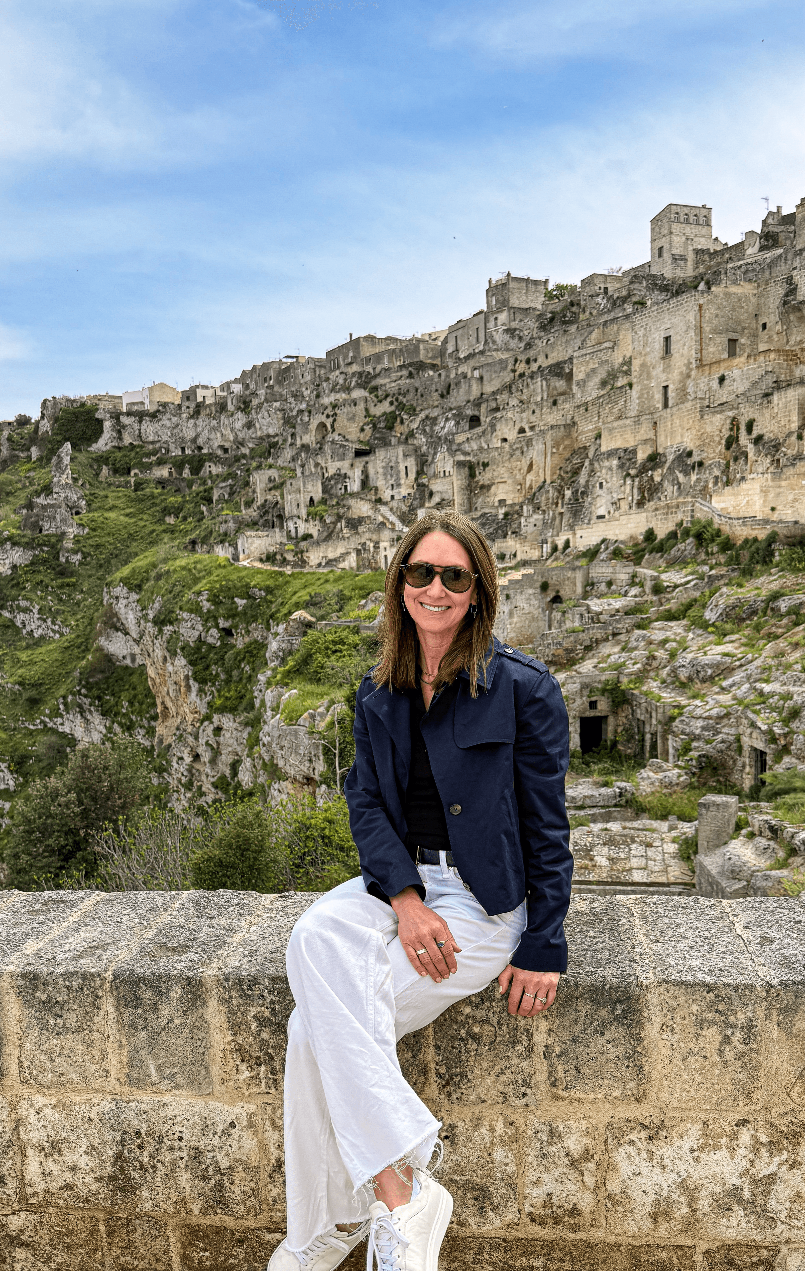 Woman sitting on a stone wall with ancient hillside buildings in the background, wearing sunglasses, a dark jacket, and white pants.