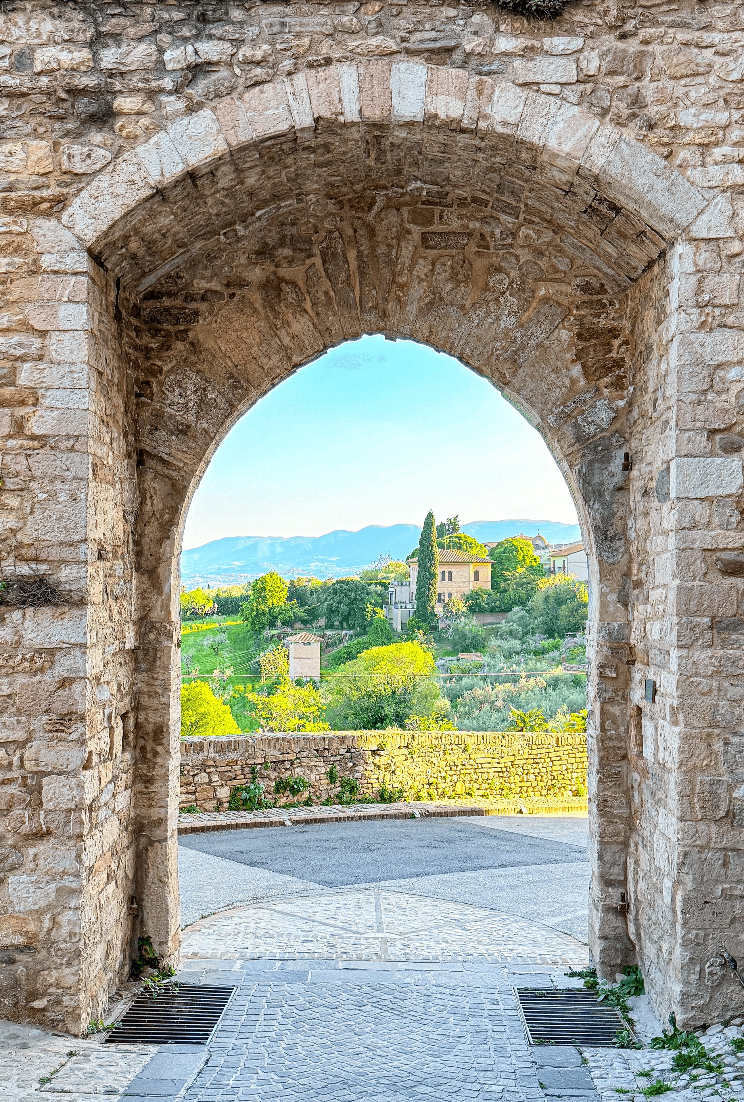 Stone archway opening to a scenic landscape with trees, a building, and distant mountains under a blue sky.