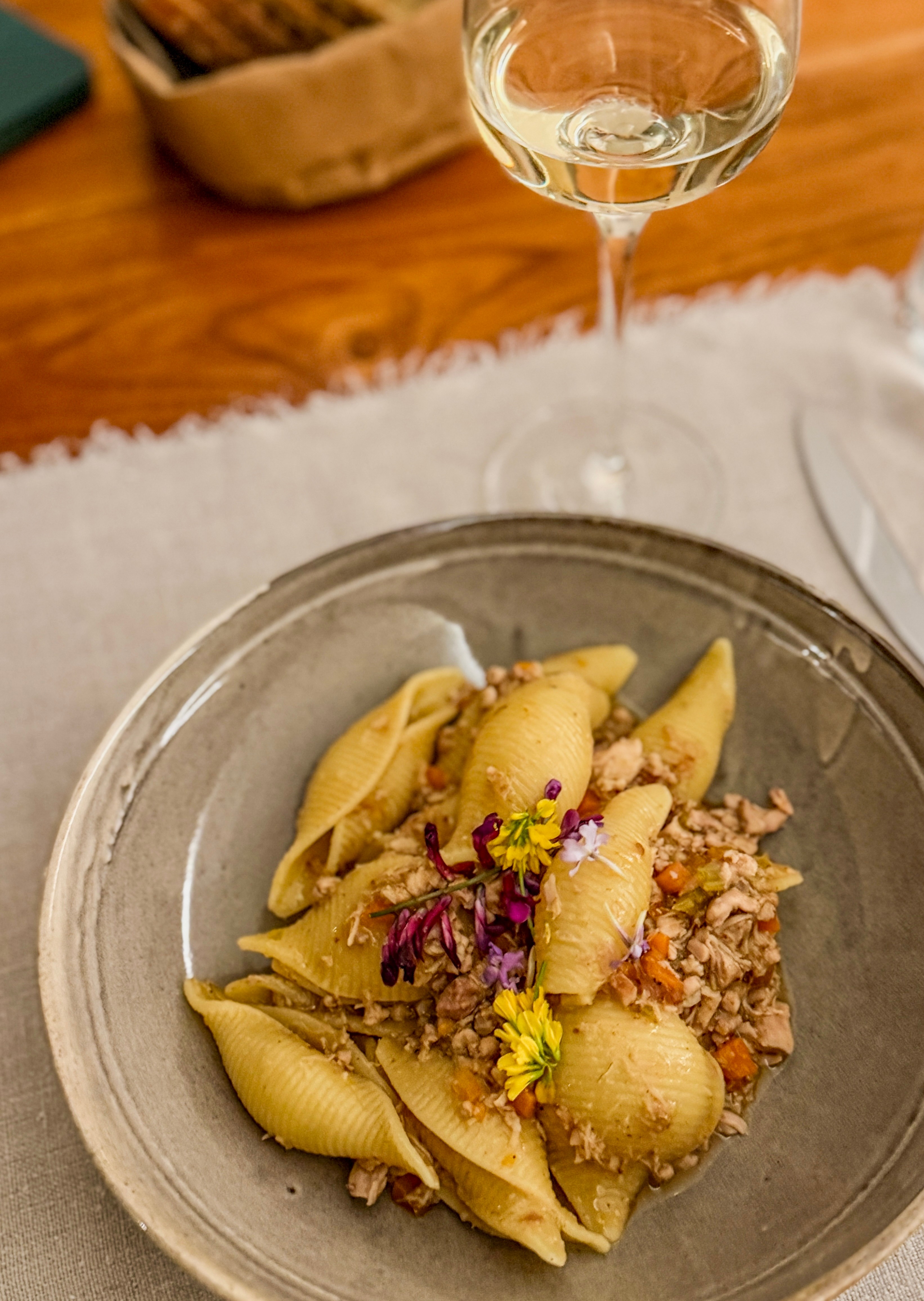 Pasta with meat, garnished with purple and yellow edible flowers, on a gray plate, with a glass of white wine in the background.