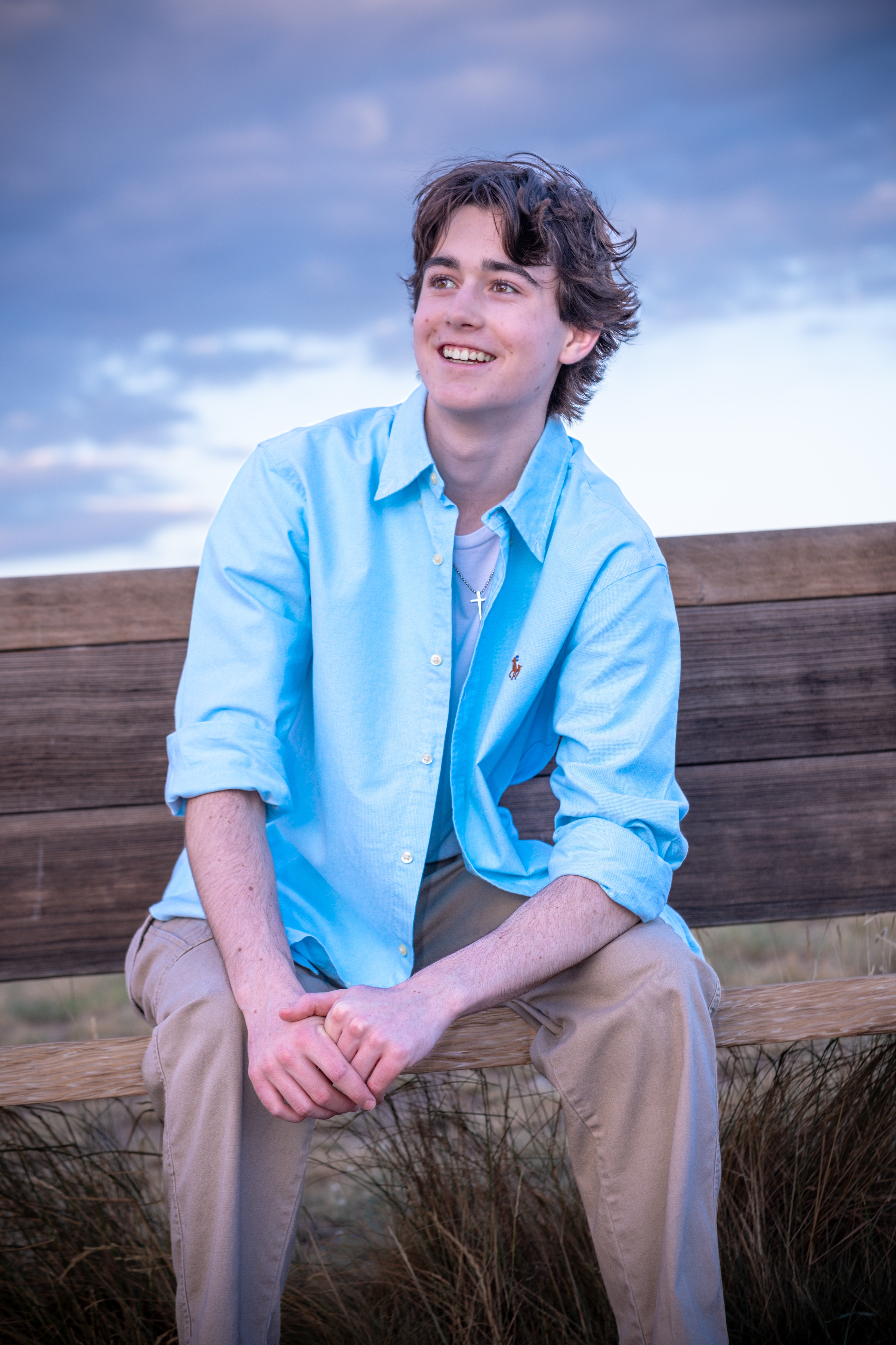 Young person with curly hair smiling, sitting outdoors on a wooden bench, wearing a light blue shirt and beige pants.