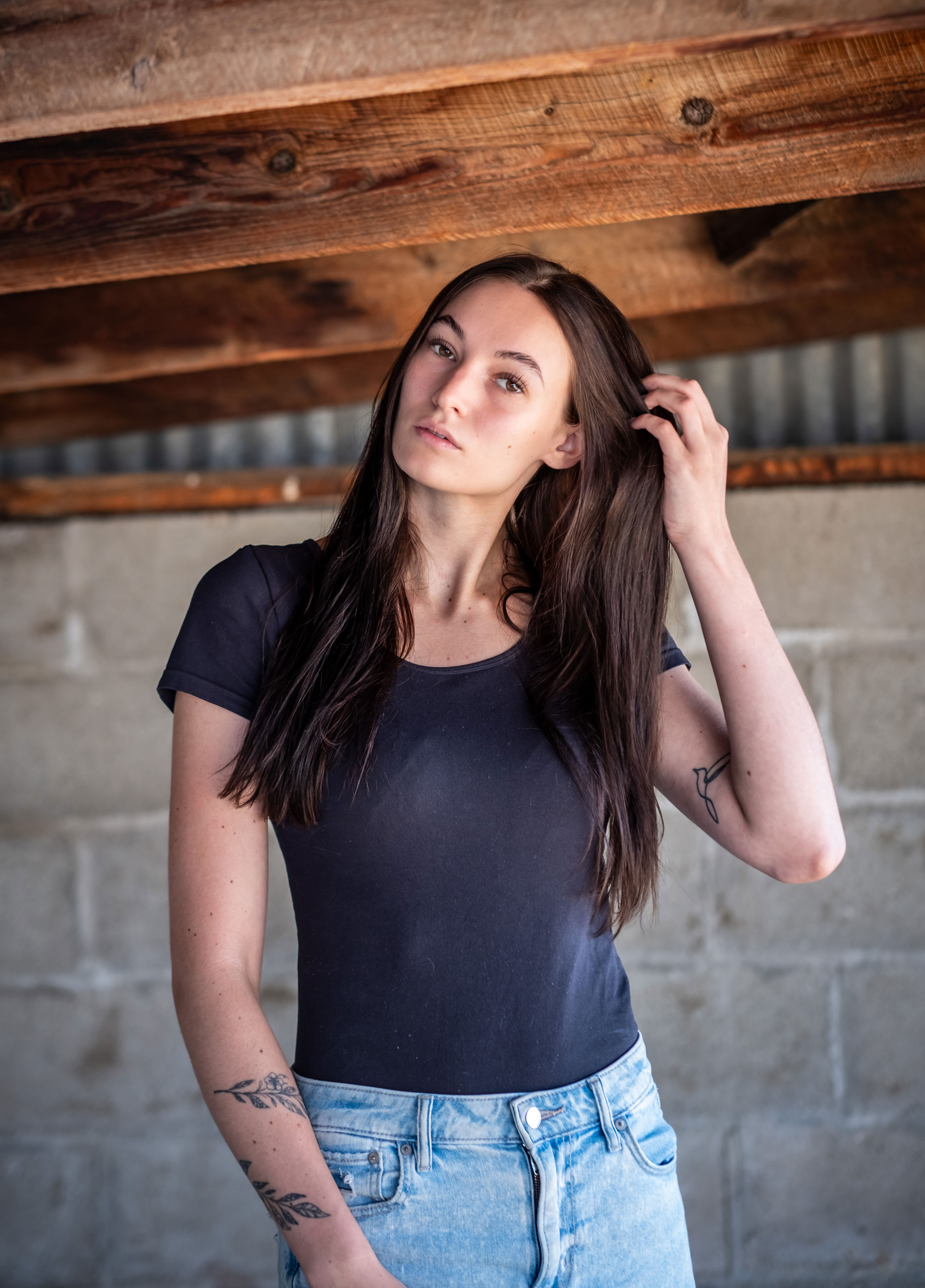 Young woman with long dark hair, wearing a black t-shirt and jeans, standing indoors with a wooden ceiling above.