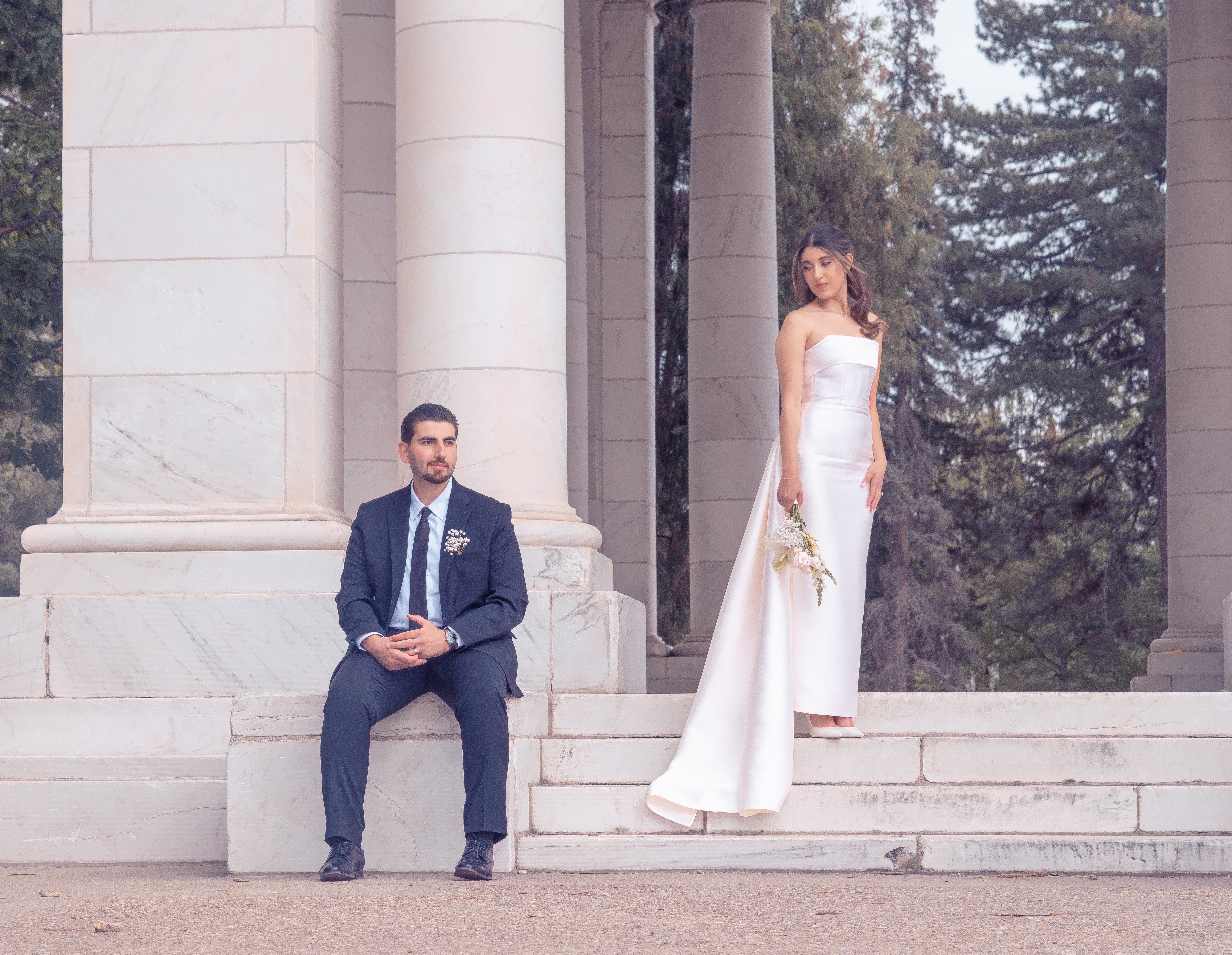 Man in a suit sitting on steps, woman in a white dress standing nearby, columns and trees in background.