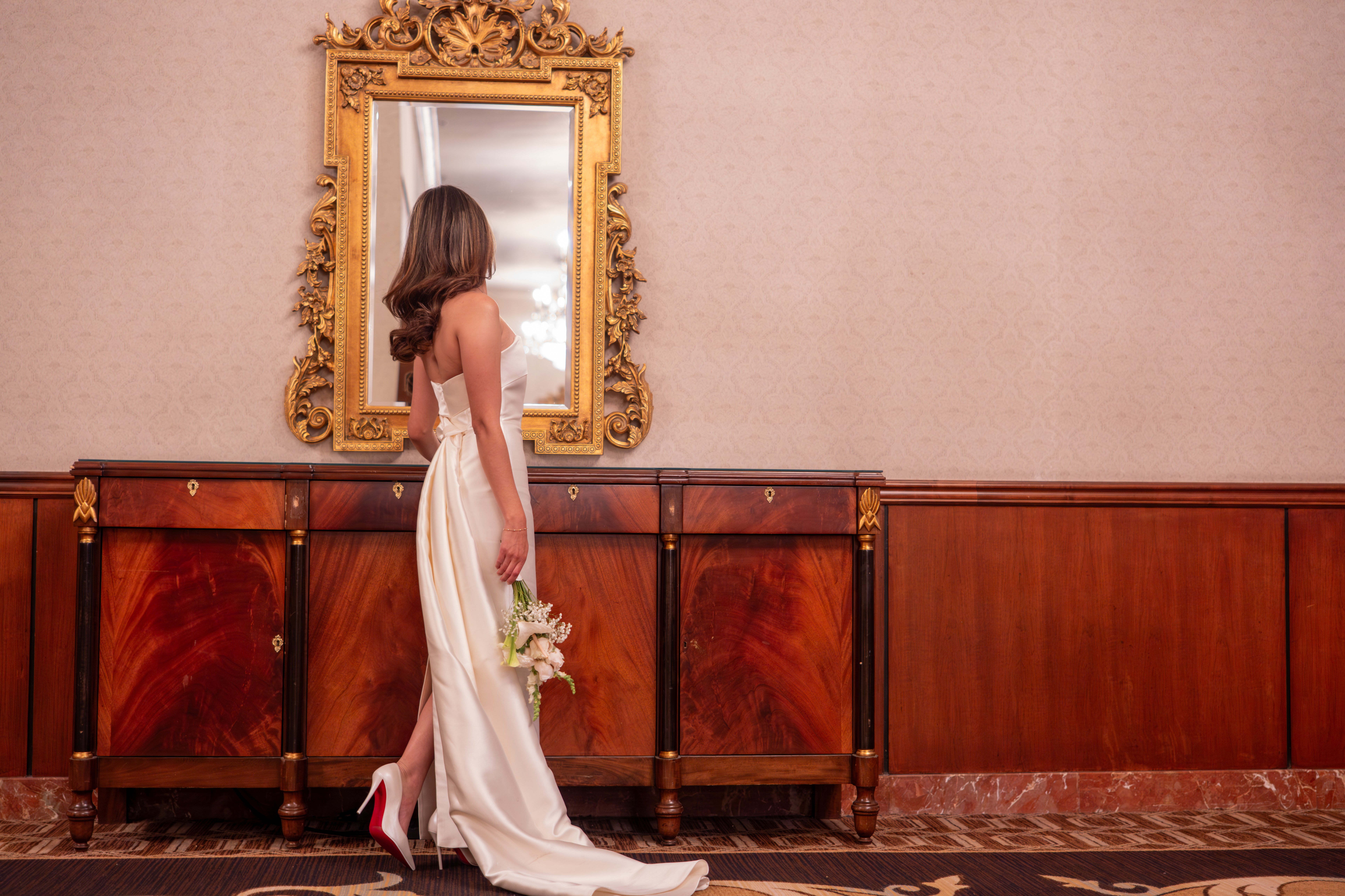 Woman in a white dress holding a bouquet, standing in front of a mirror with a gold frame, in a decorated room.