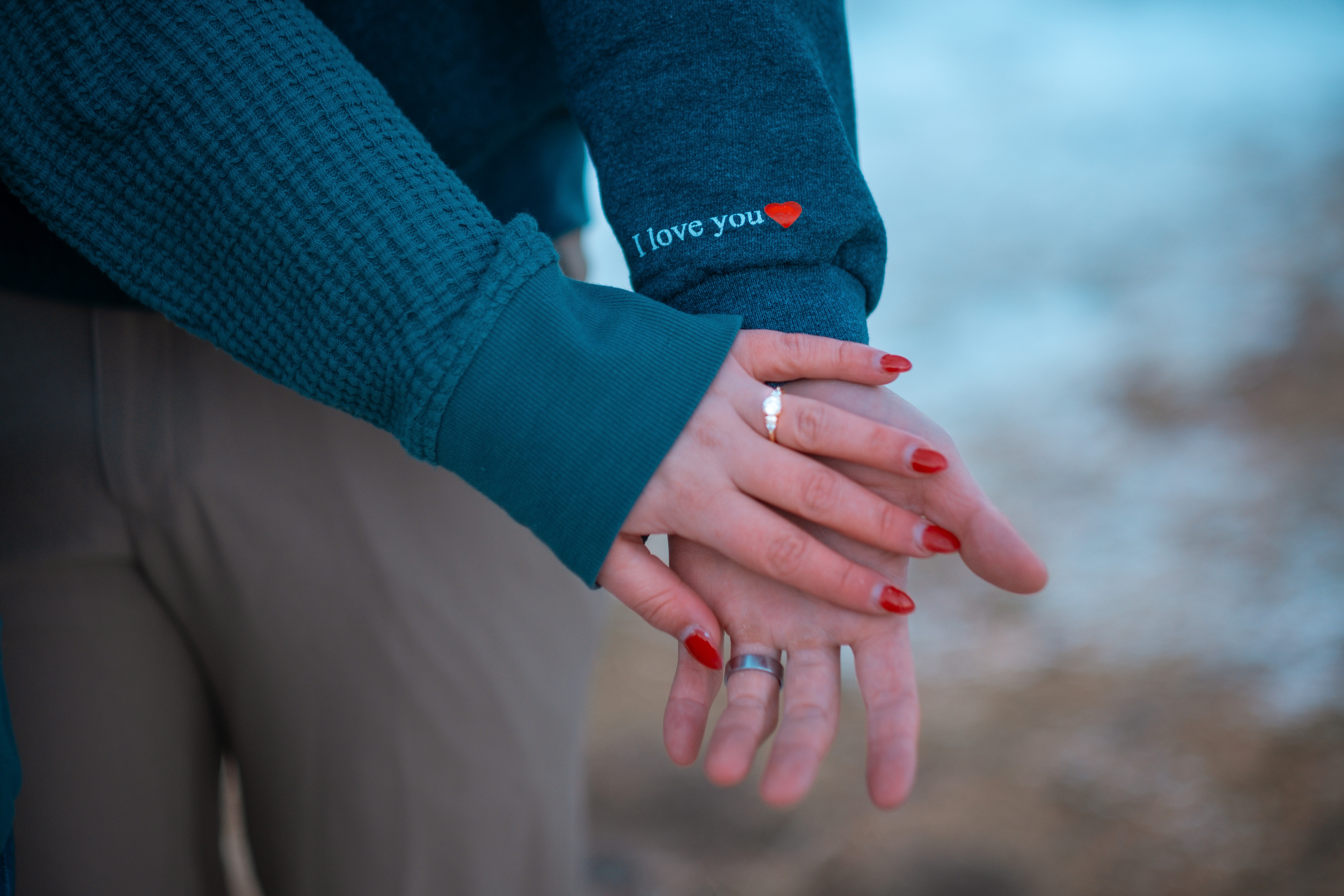 Two people hold hands, one with a ring, outdoors with blurred background, one person wearing a blue sweater, the other a dark jacket with a small red heart and text.