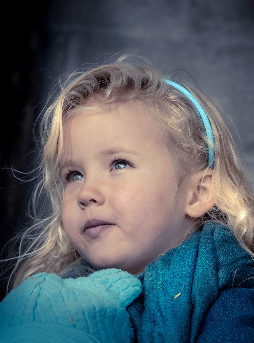 Young girl with blonde hair and blue headband looking upward, wearing a blue sweater, outdoors.