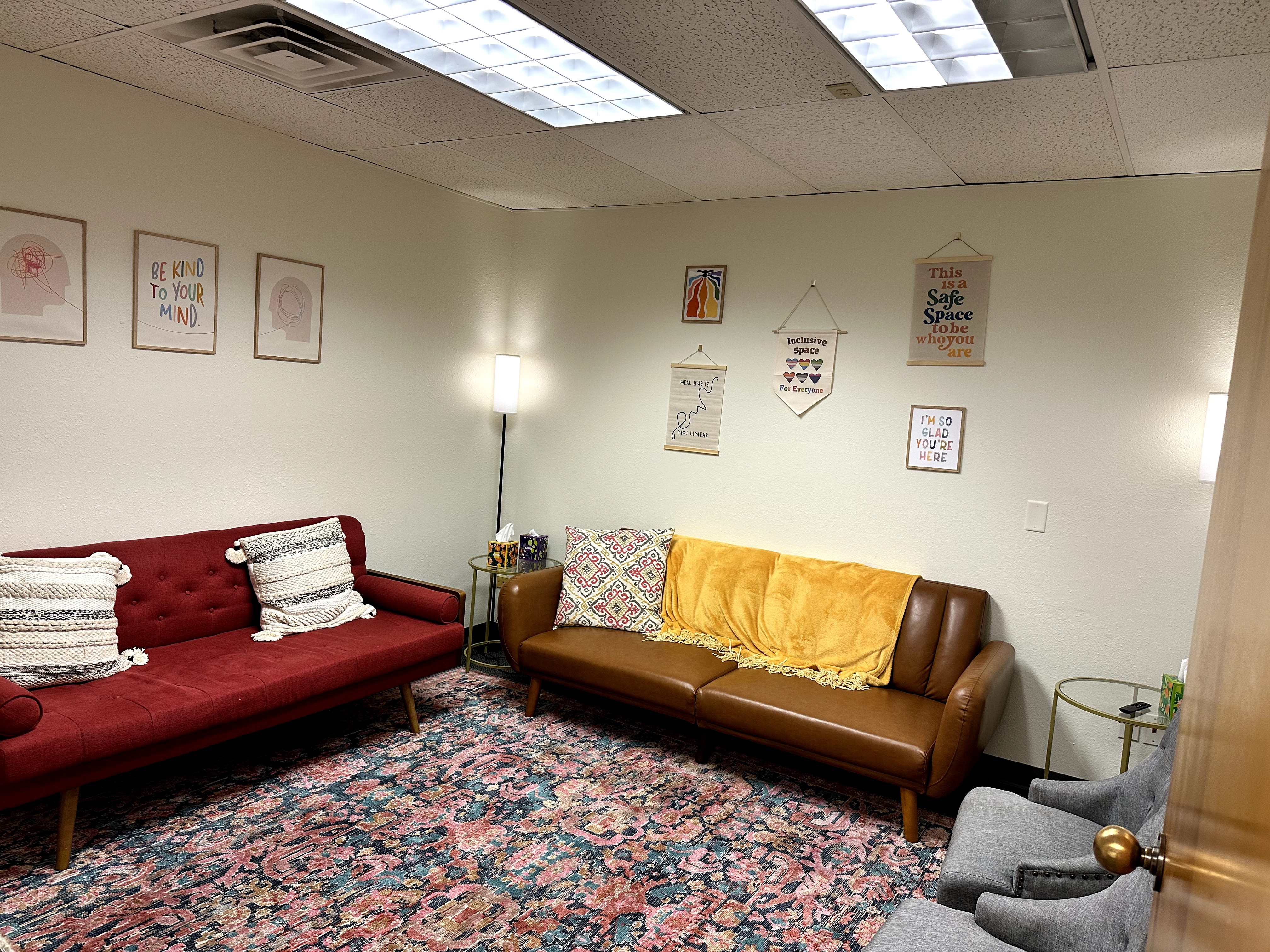 Living room with two sofas, one red and one brown, a patterned rug, wall art, and a lamp, arranged in a cozy setting.