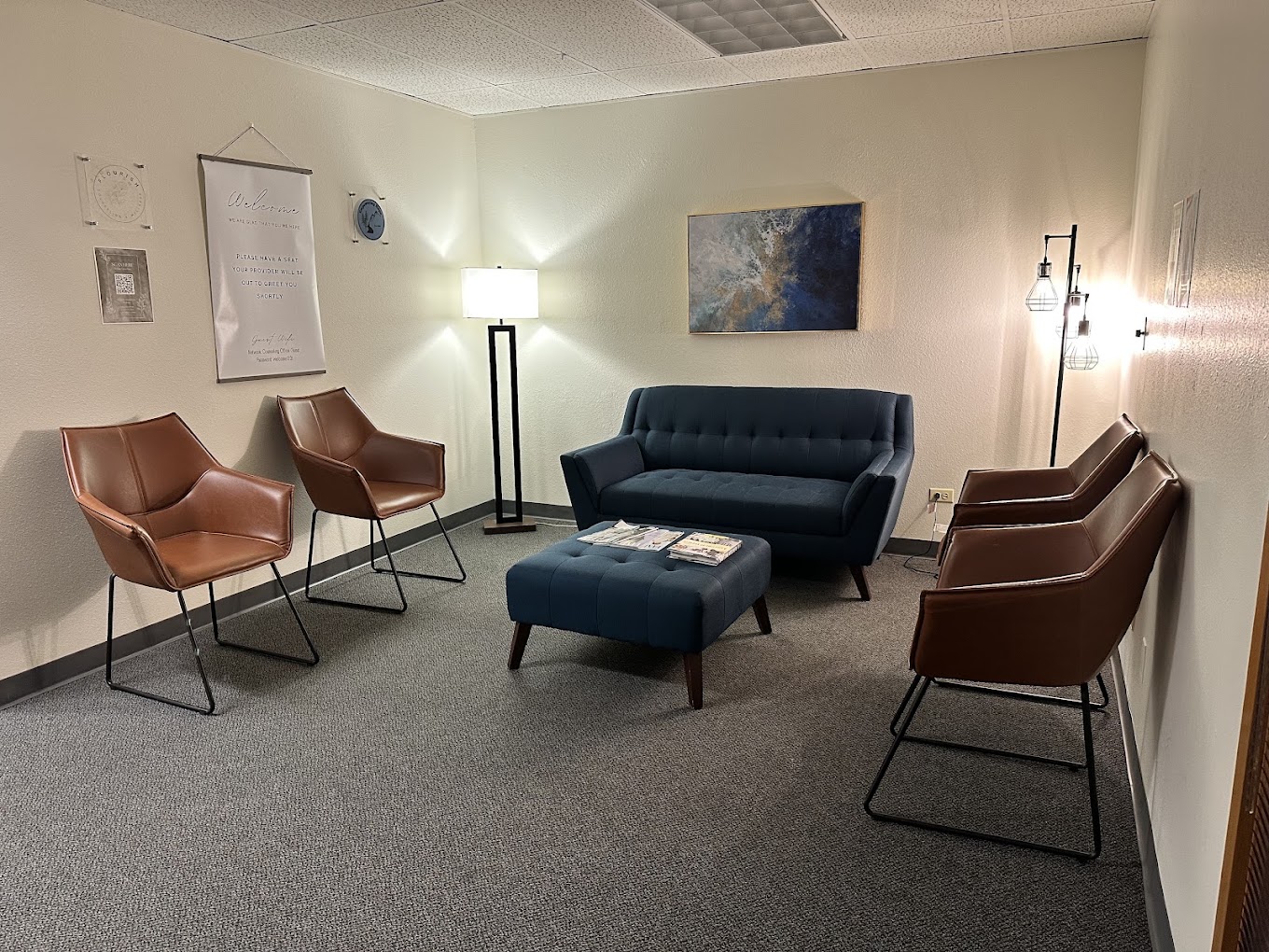 Waiting room with a blue sofa, four brown chairs, a floor lamp, and a painting on the wall.