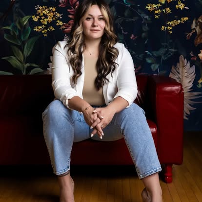 Young woman with long wavy hair sitting on a red sofa, wearing a white blazer and jeans, in front of a floral background.