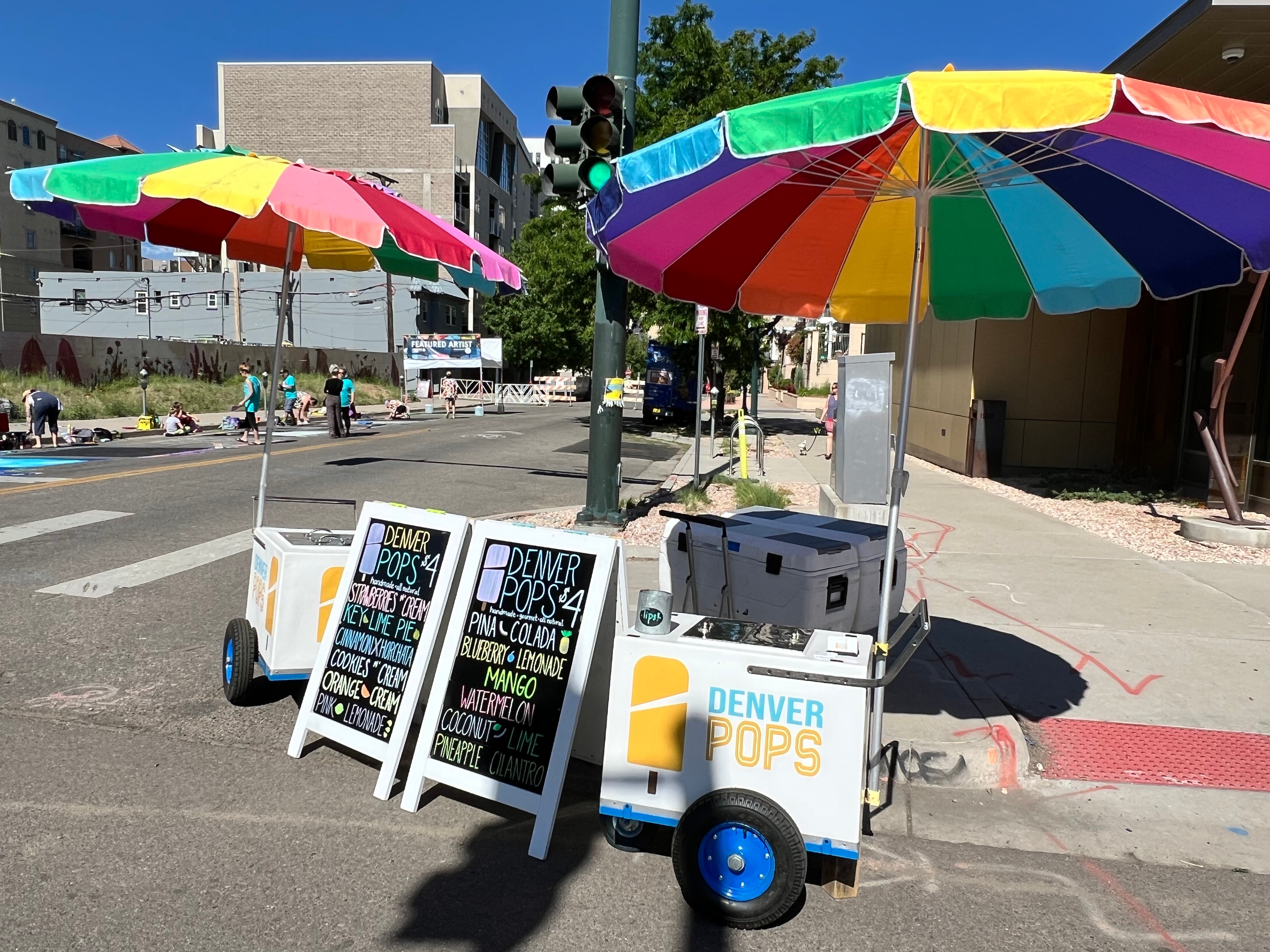 Street vendor cart with colorful umbrellas and chalkboard signs on sidewalk, city buildings in background.