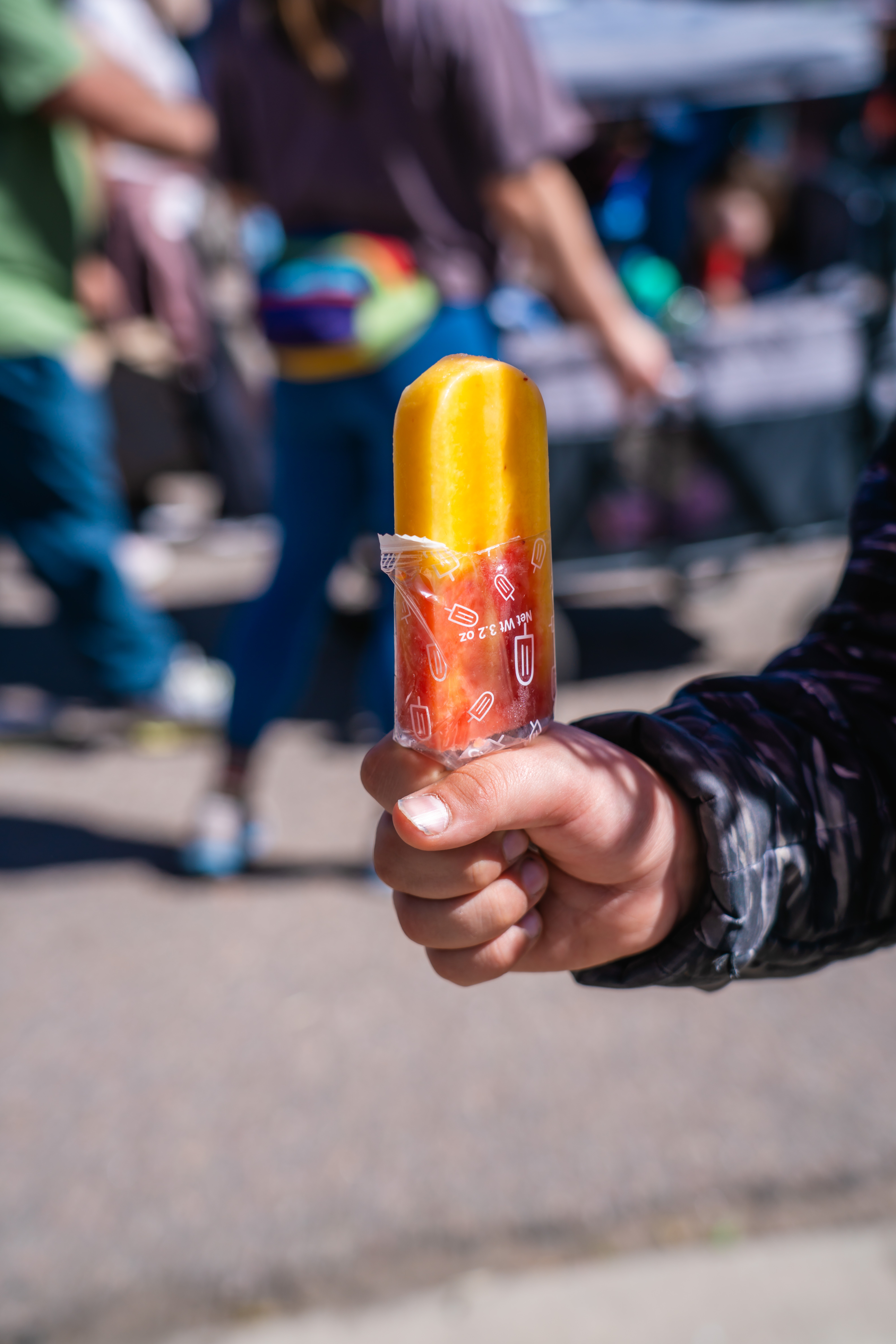 Person holding a yellow and red popsicle in a busy outdoor setting with people in the background.